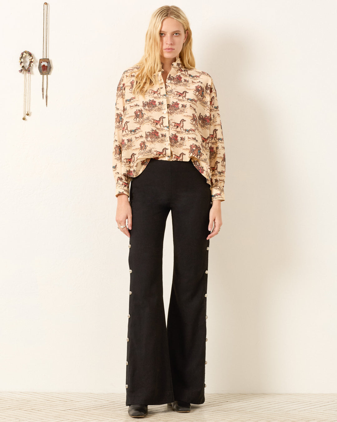 A woman stands indoors wearing the Kiki Canyon Cowboy Shirt and high-waisted black flared pants with side buttons. Two decorative objects hang on the wall to her left.