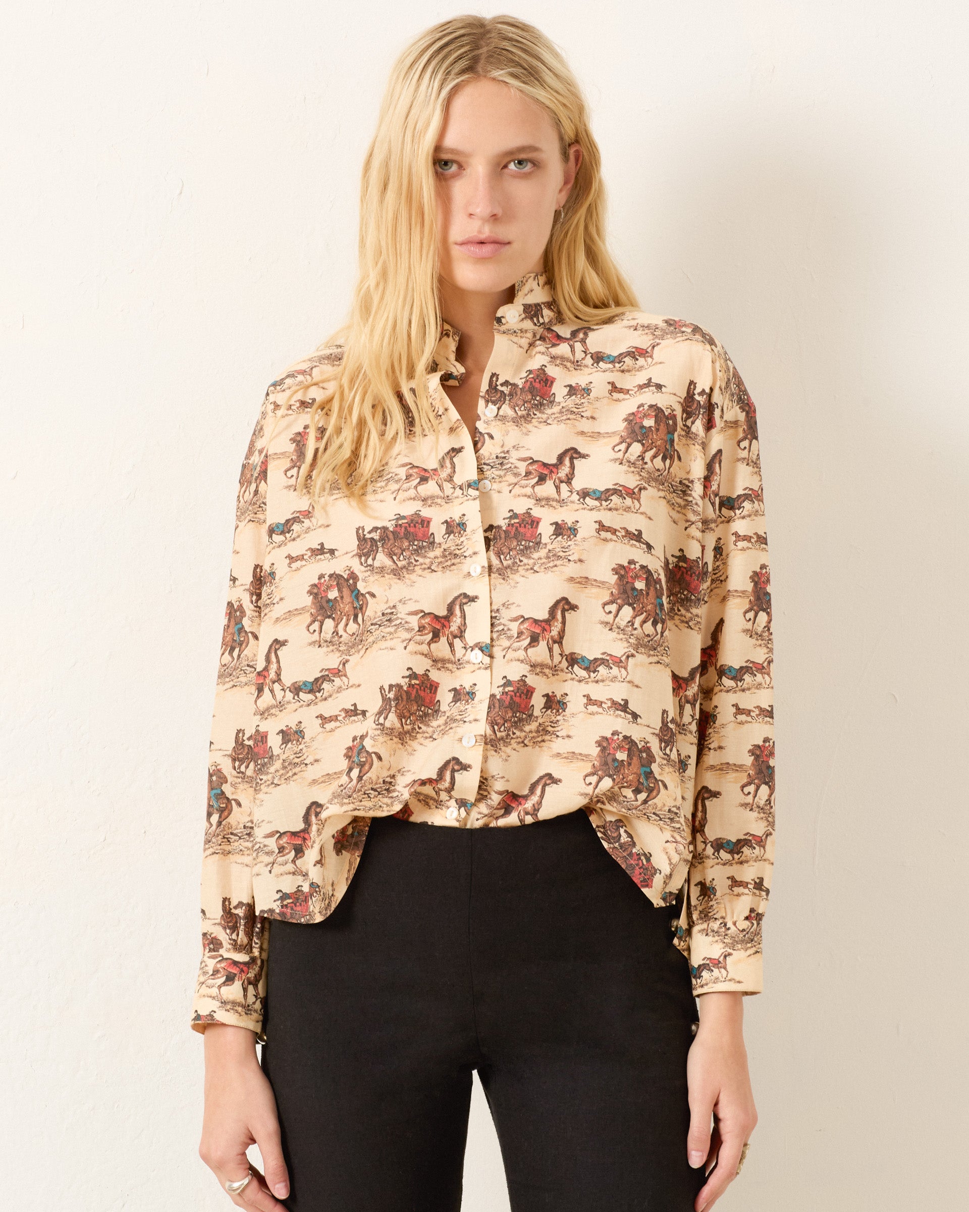 A woman with long blonde hair wears the Kiki Canyon Cowboy Shirt, a beige cotton-silk blend with a horse and rider print, paired with high-waisted black pants, standing against a plain light-colored wall.