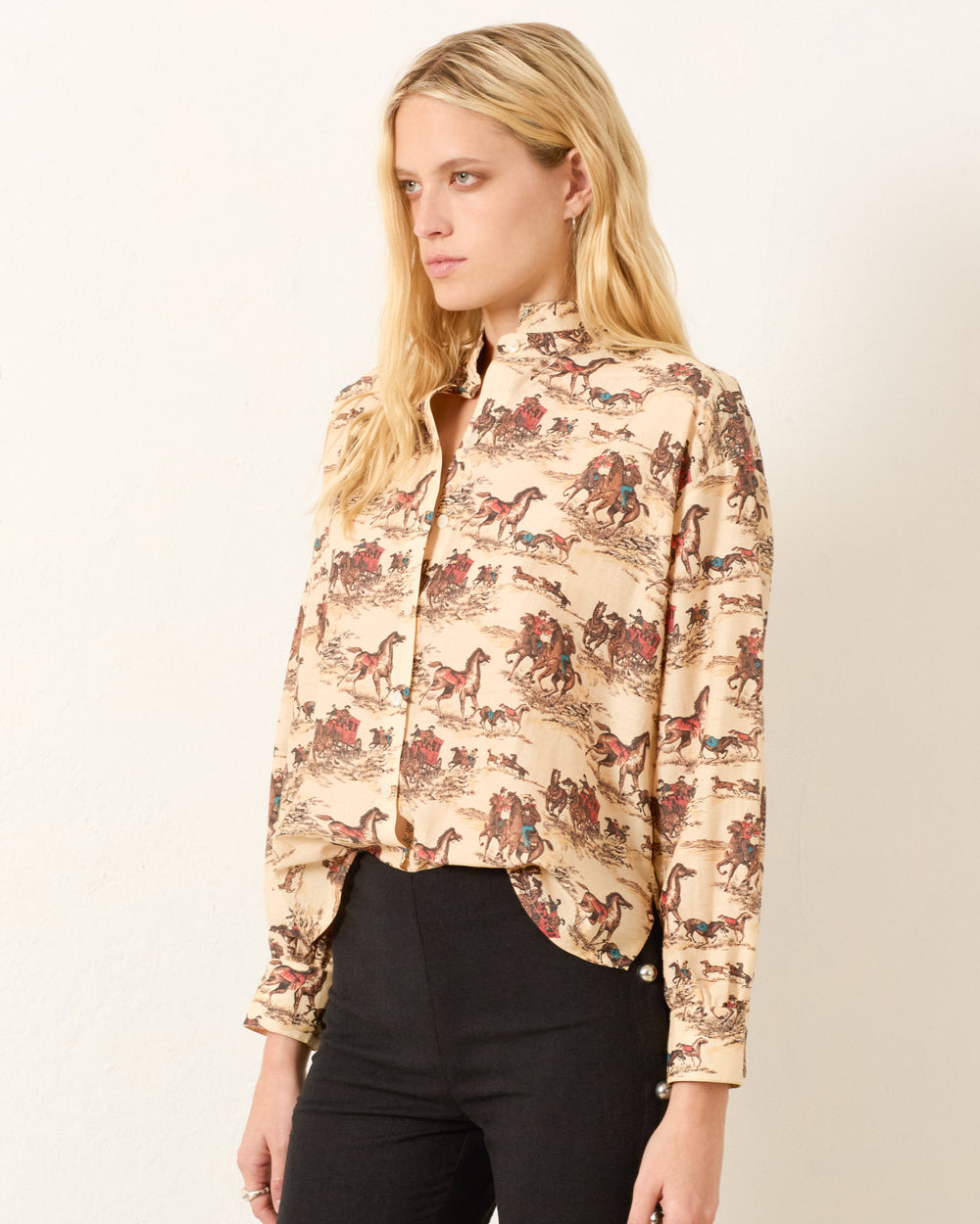 A woman with long blonde hair wears the Kiki Canyon Cowboy Shirt, a high-neck, button-up Edwardian style in a cotton-silk blend with an equestrian print, paired with black pants against a plain light background.