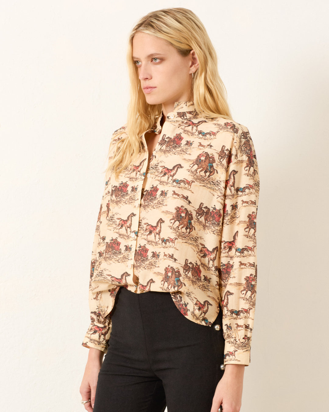 A woman with long blonde hair wears the Kiki Canyon Cowboy Shirt, a high-neck, button-up Edwardian style in a cotton-silk blend with an equestrian print, paired with black pants against a plain light background.