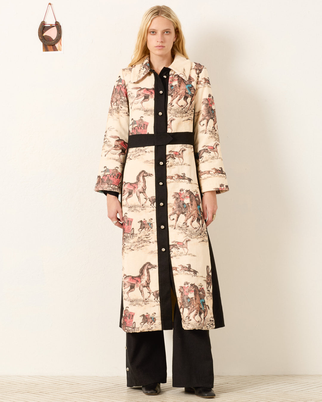 A woman stands against a white wall, wearing the Lara Canyon Cowboy Coat featuring horse and rider illustrations over black pants and shoes. A small decorative item hangs on the wall beside her.