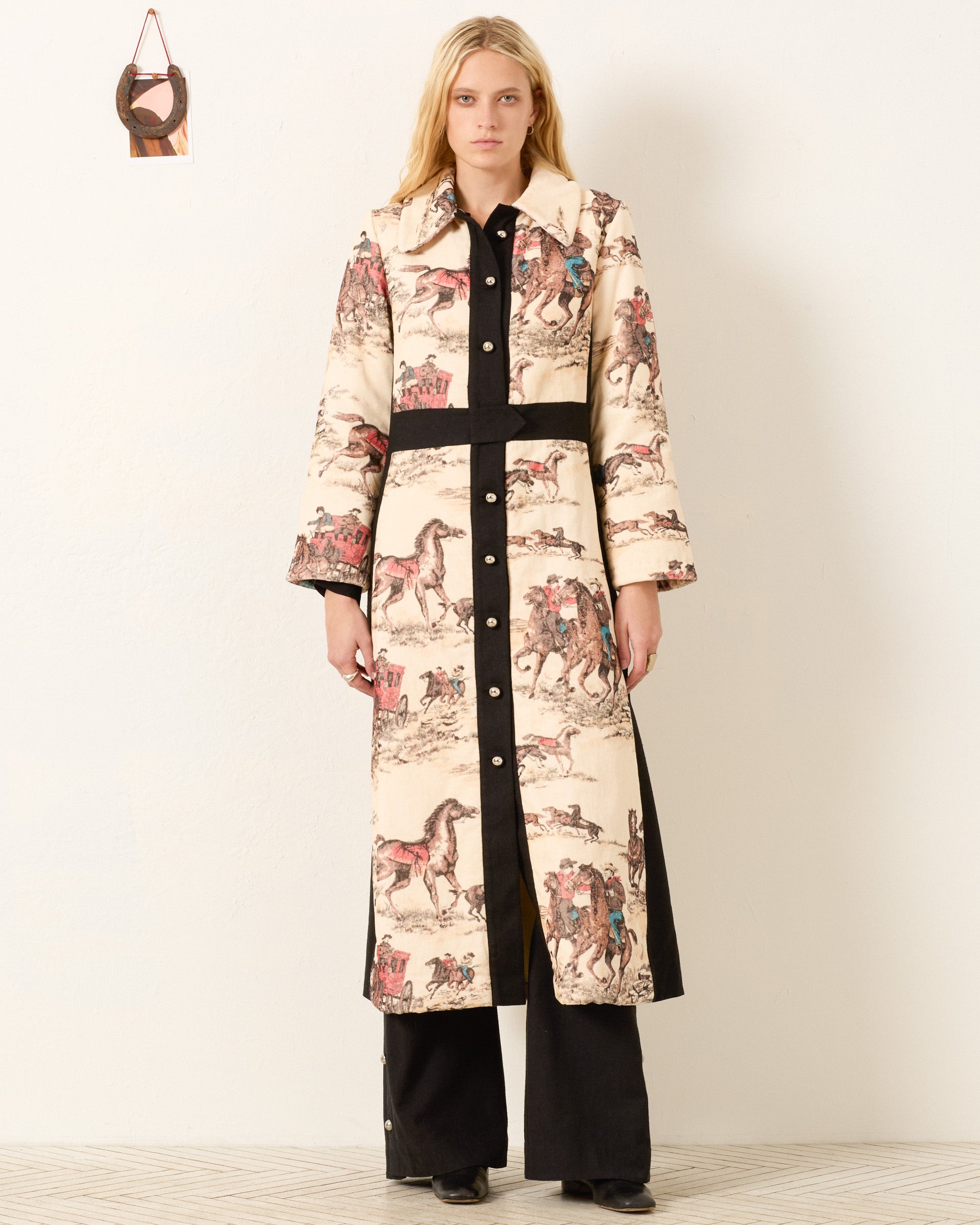 A woman stands against a white wall, wearing the Lara Canyon Cowboy Coat featuring horse and rider illustrations over black pants and shoes. A small decorative item hangs on the wall beside her.