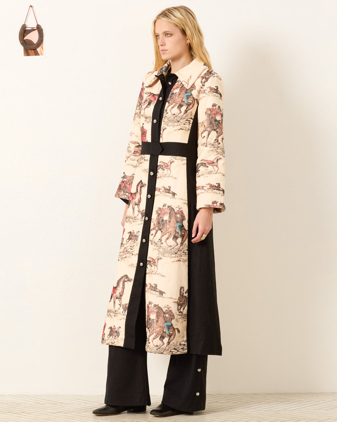 A woman stands indoors facing sideways, wearing the Lara Canyon Cowboy Coat with horse prints and black side panels, paired with wide-leg black pants. The vintage-inspired piece highlights Western charm against a plain wall.