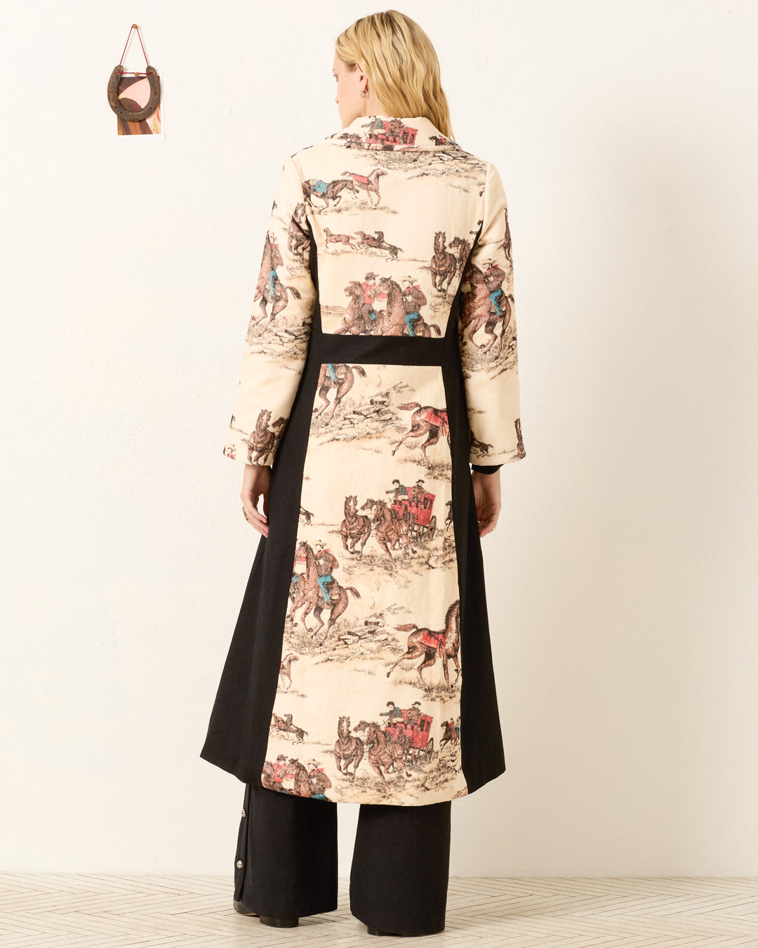 A woman faces away wearing the Lara Canyon Cowboy Coat, featuring a horse and rider print on the back and sleeves in a cowboy desert motif, paired with black pants.