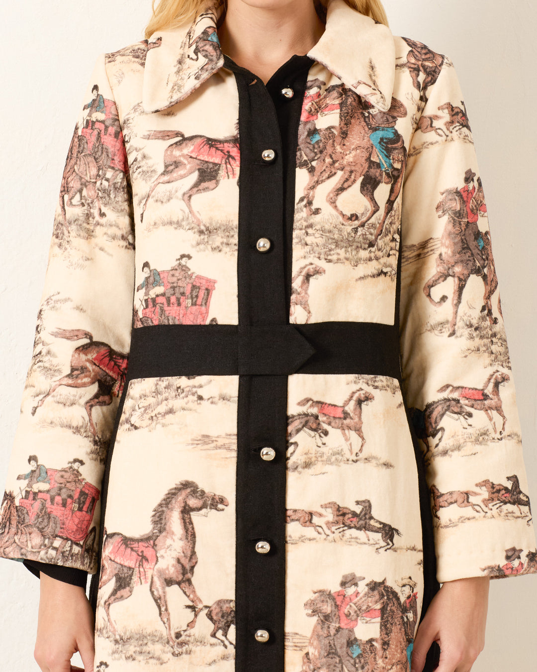 A person wearing the Lara Canyon Cowboy Coat—a long, cream cotton-velvet Western coat with black trim and a vintage print of horses, riders, and stagecoaches in muted tones.