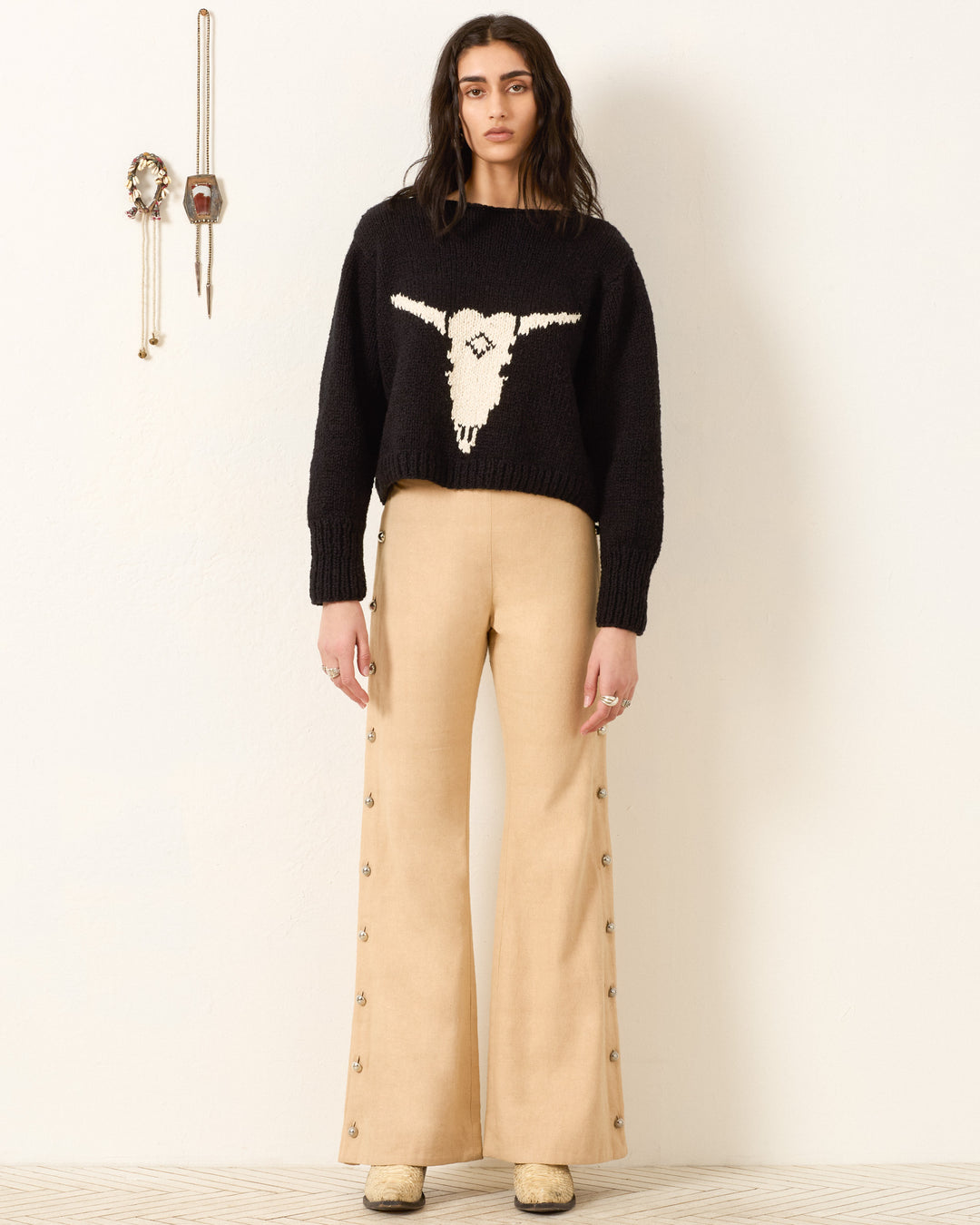 A woman stands against a white wall wearing the Leigh Cow Skull Sweater, beige flared pants with side buttons, and cream shoes. Southwestern jewelry decorates the wall behind her.