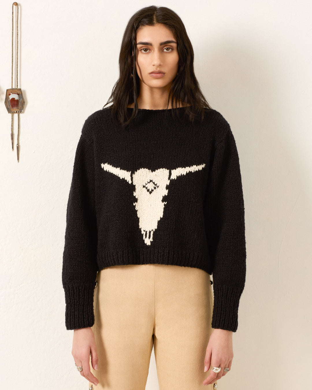 A person with long dark hair wears the Leigh Cow Skull Sweater in black, paired with beige pants, standing against a plain wall with a small hanging object.