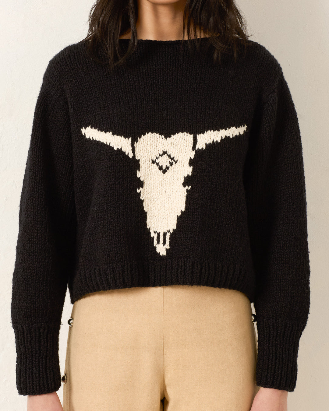 A person wearing the black Leigh Cow Skull Sweater, featuring a white bull skull design on the front, paired with beige pants.