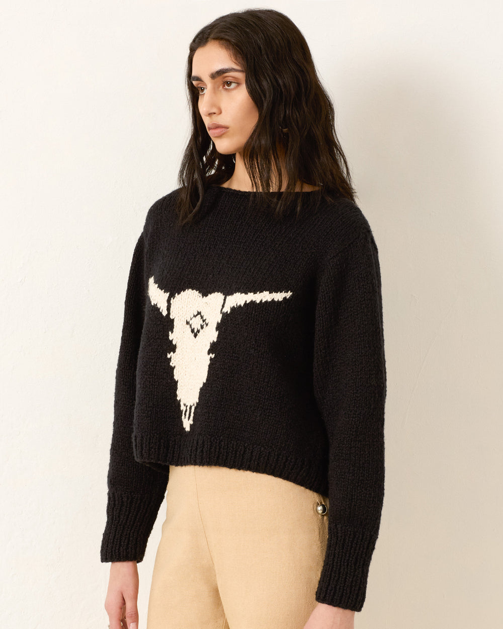 A woman wears the Leigh Cow Skull Sweater in black, featuring a bold white graphic on the front, paired with beige pants and standing against a plain light-colored wall.