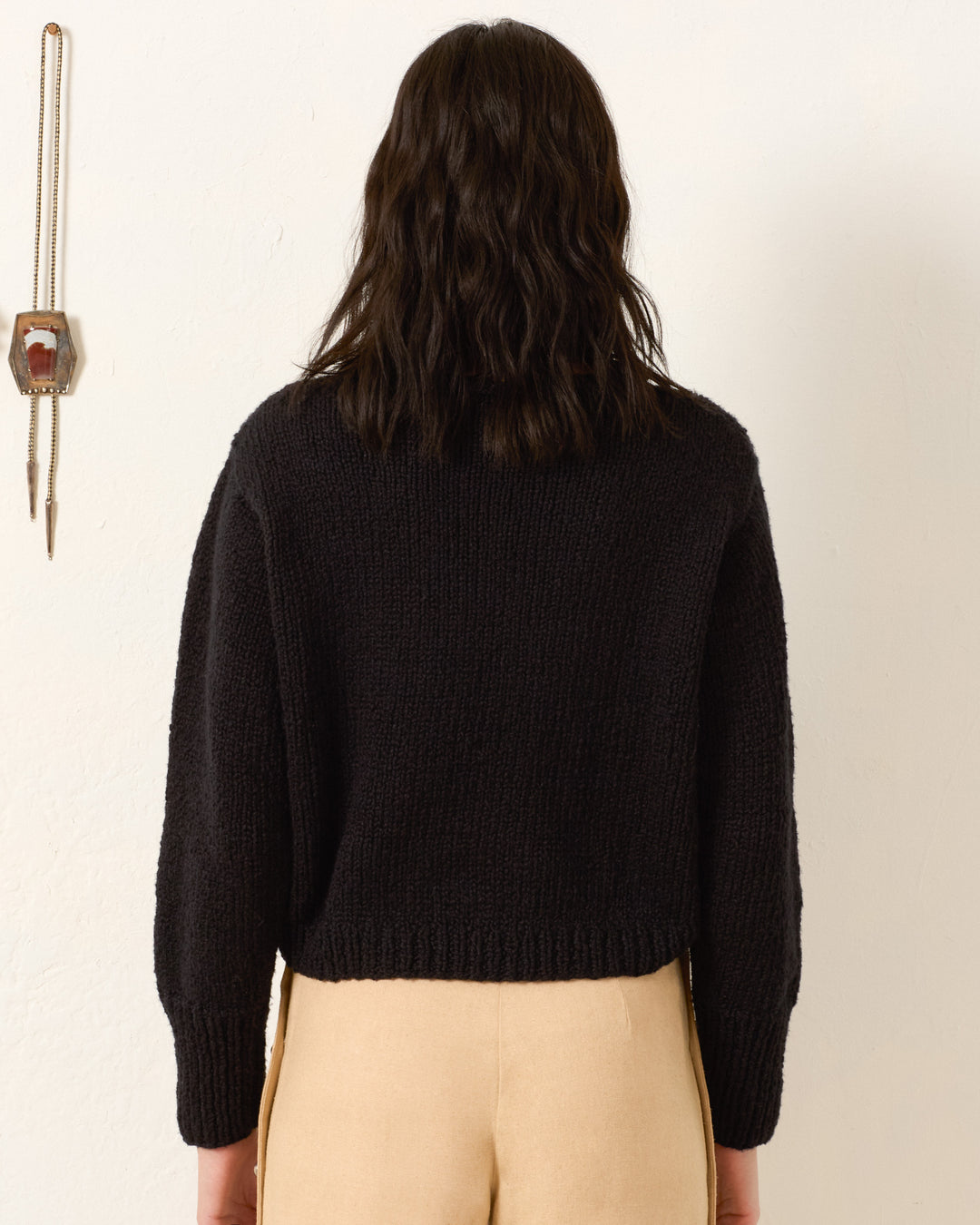 A person with shoulder-length dark hair, wearing the black Leigh Cow Skull Sweater and beige pants, stands with their back to the camera against a plain white wall.