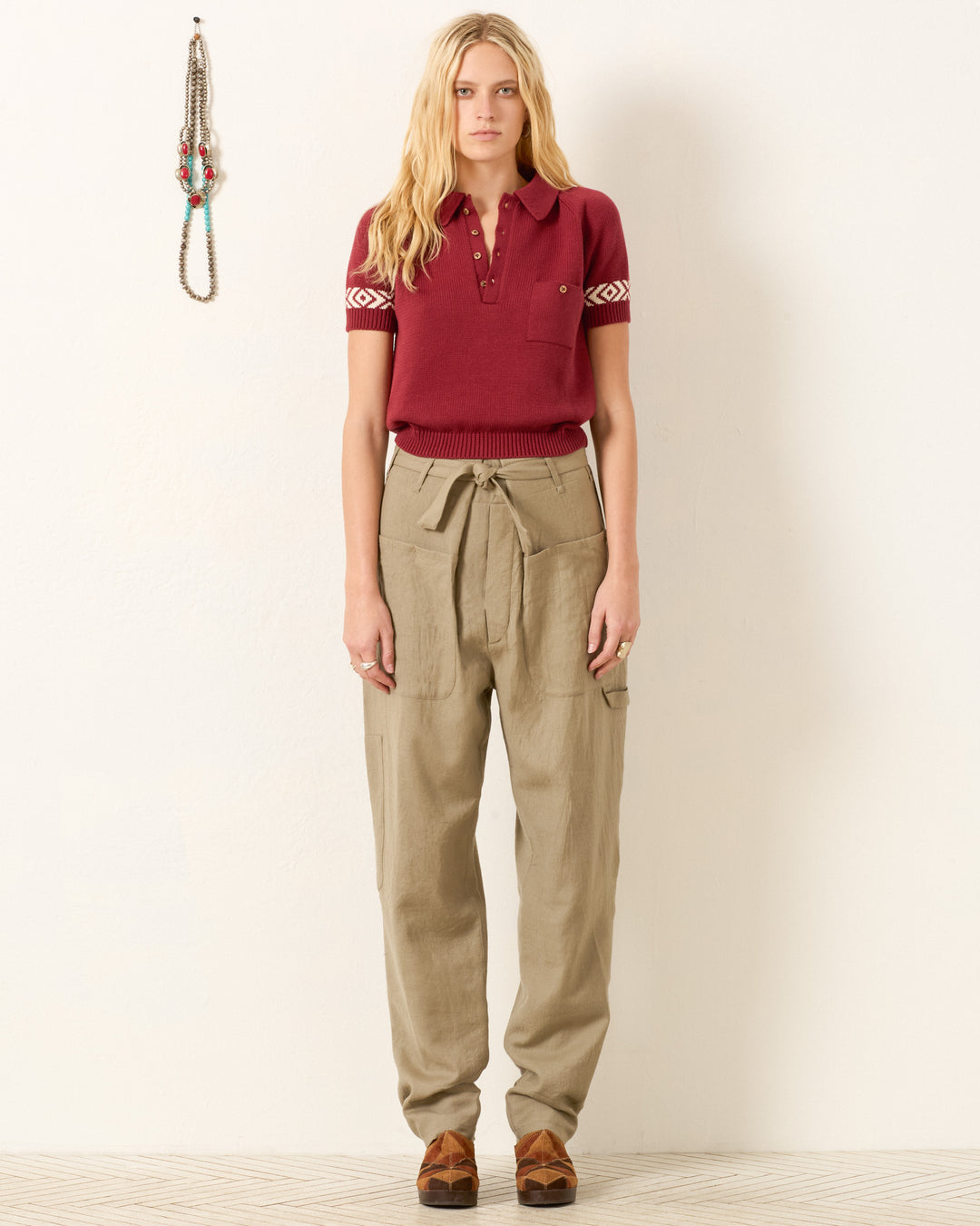 A person stands facing forward against a white wall, wearing the Lessi Mesa Knit Top in maroon and high-waisted beige trousers with a tie belt. Necklaces hang on the wall to the left.