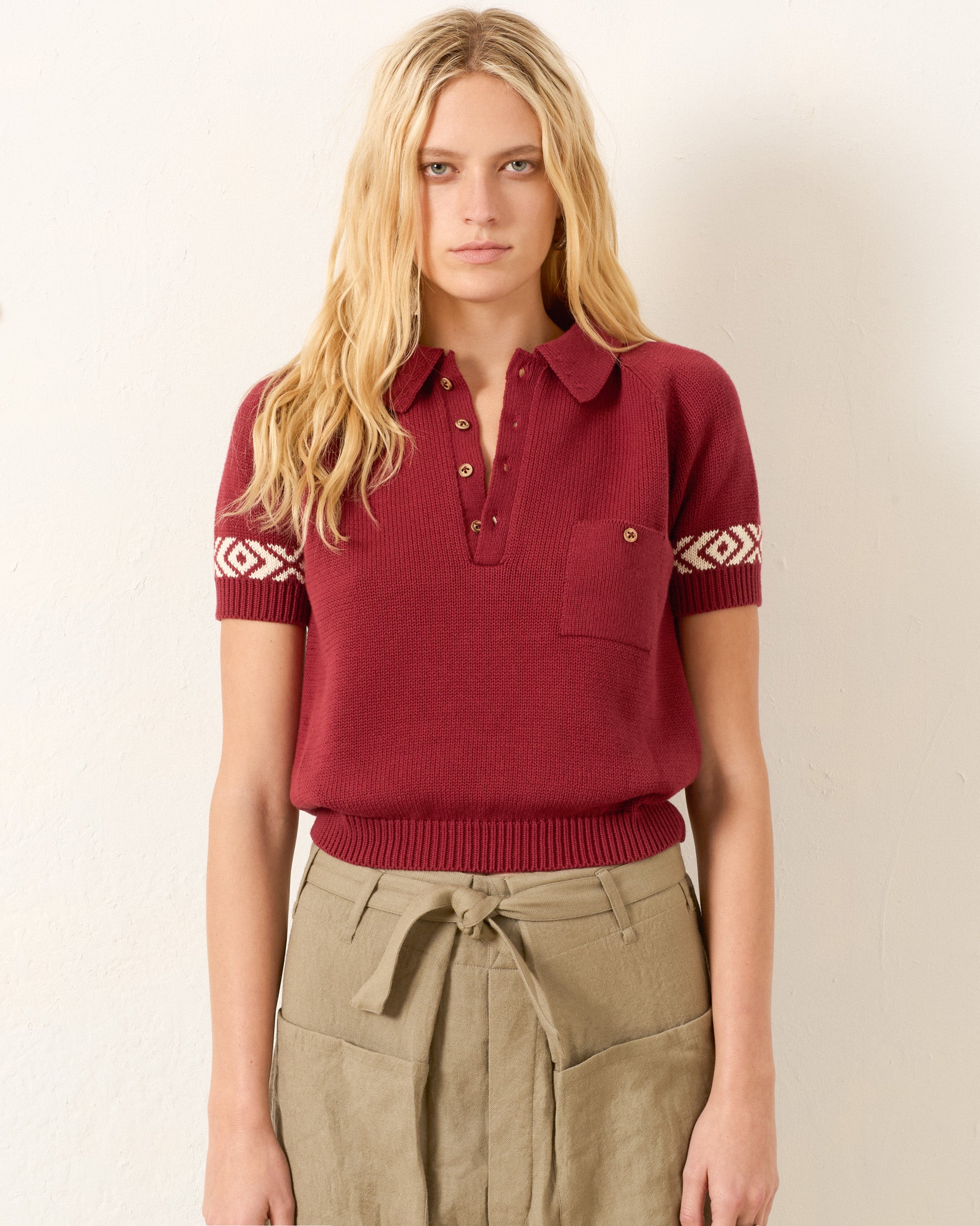 A person with long blonde hair wears the Lessi Mesa Knit Top, a cropped maroon polo sweater with buttons and a chest pocket, styled with high-waisted belted khaki pants, against a plain background.