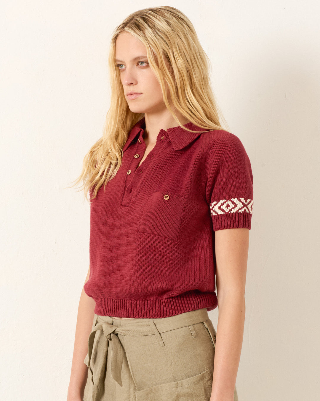 A woman with long blonde hair wears the Lessi Mesa Knit Top, a cropped maroon cotton polo sweater with a chest pocket and patterned sleeves, paired with high-waisted beige pants.