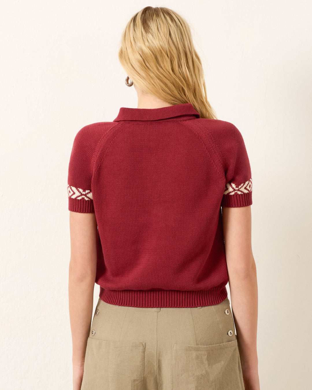 A person with long blonde hair is shown from the back wearing the Lessi Mesa Knit Top, a cropped maroon sweater with patterned sleeve details, paired with a tan skirt.