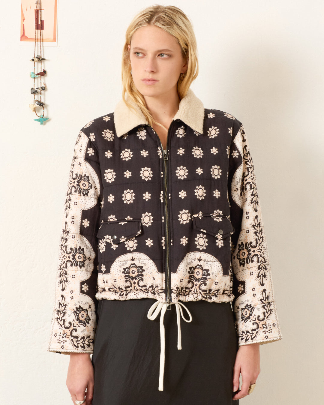 A woman stands against a plain wall wearing the Lex Bronco Bandana Jacket, featuring a black and white bandana print, wool sherpa collar, and drawstring waist.