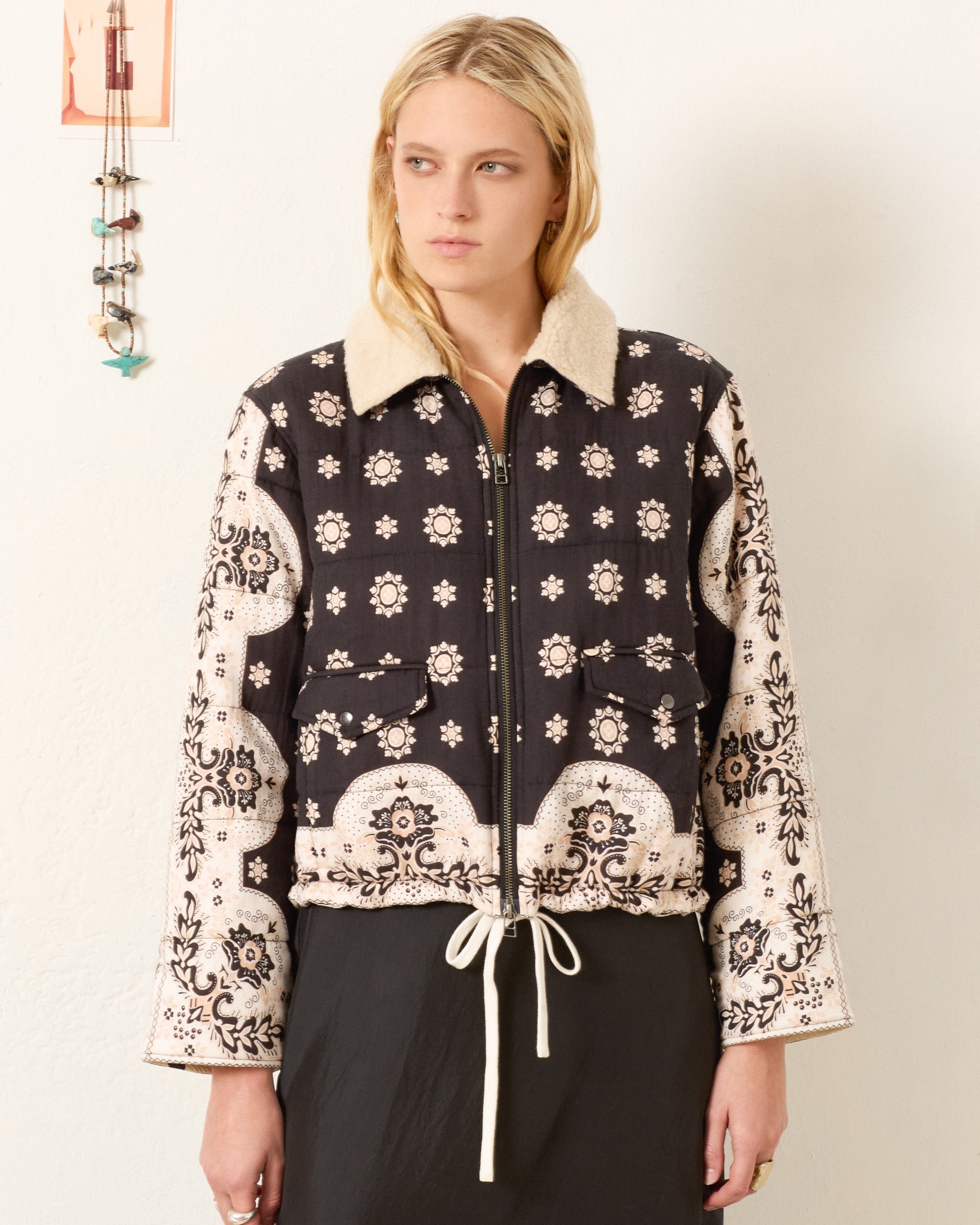 A woman stands against a plain wall wearing the Lex Bronco Bandana Jacket, featuring a black and white bandana print, wool sherpa collar, and drawstring waist.