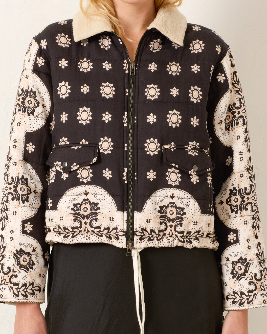 A woman wears the Lex Bronco Bandana Jacket—a black and beige bandana-print style with a zipper, two front pockets, and a wool sherpa collar. Only her torso is visible.