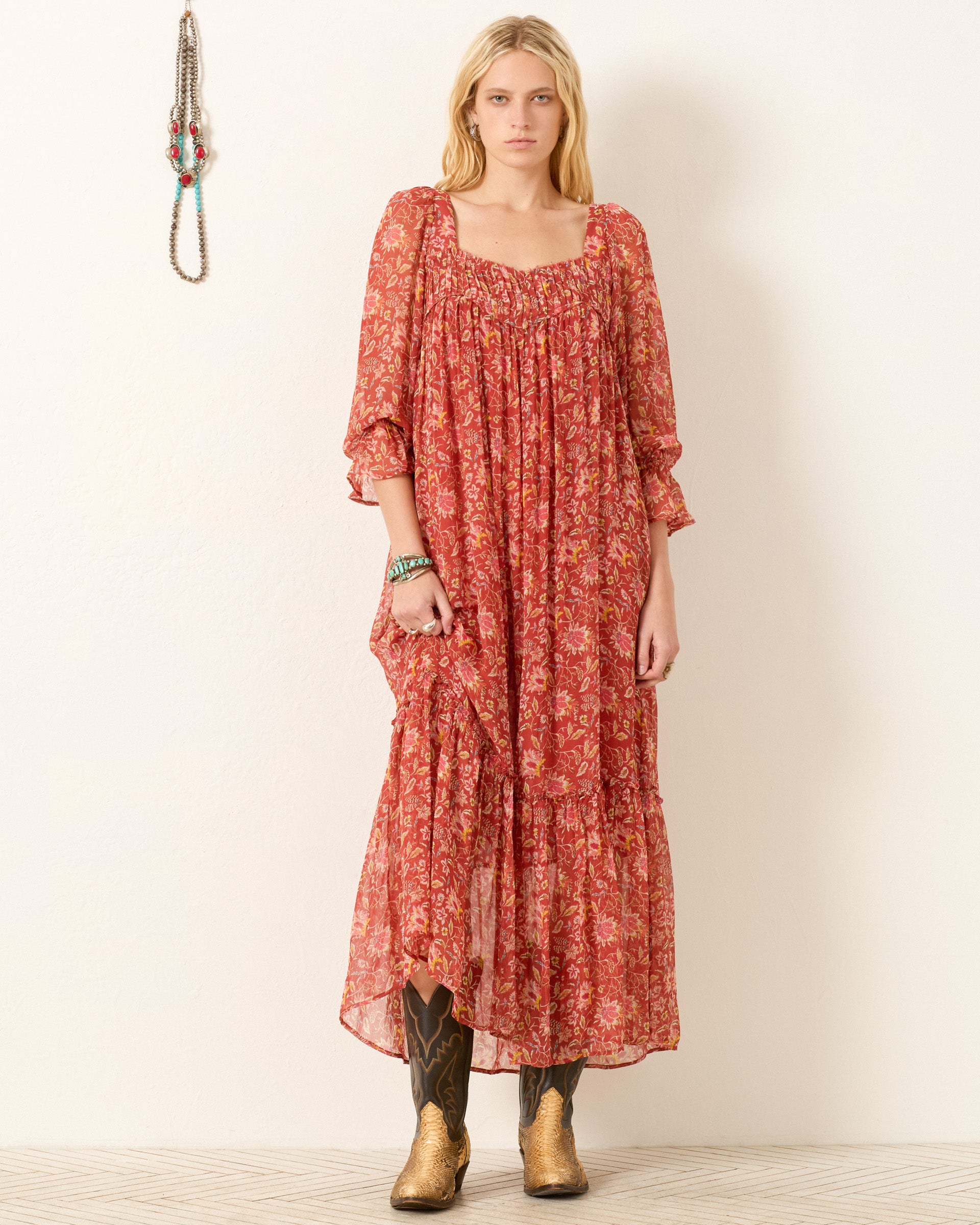 A woman with long blonde hair stands indoors wearing the Lilium Sunset Ember Dress, a floral maxi with a flowy silhouette. She pairs it with gold cowboy boots and minimal accessories. A necklace hangs on the wall beside her.