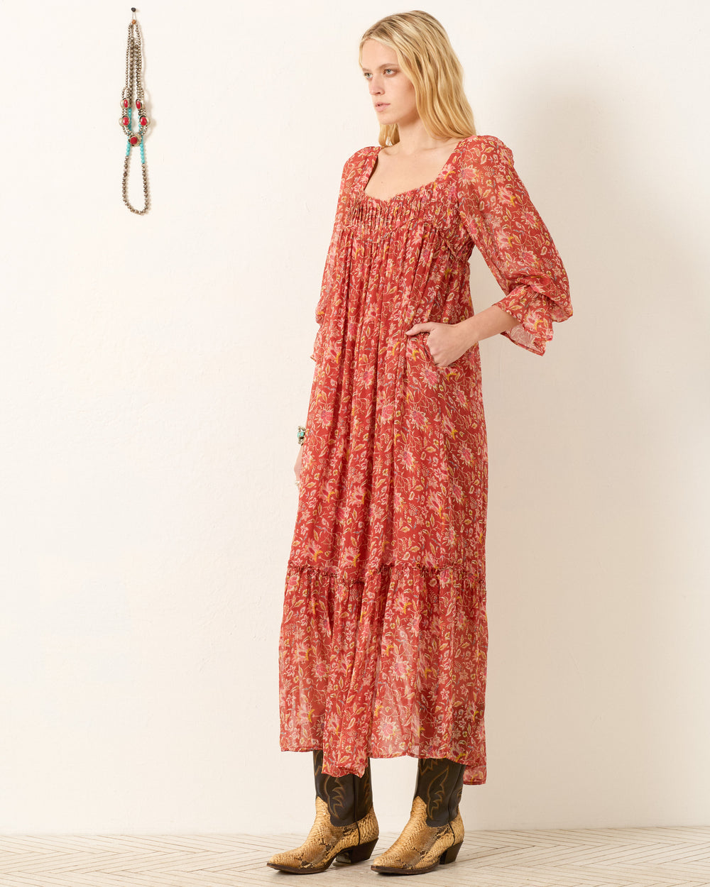 A woman in the Lilium Sunset Ember Dress, a red floral maxi with a flowy silhouette, pairs it with brown cowboy boots and stands sideways by a plain light wall, with necklaces displayed beside her.