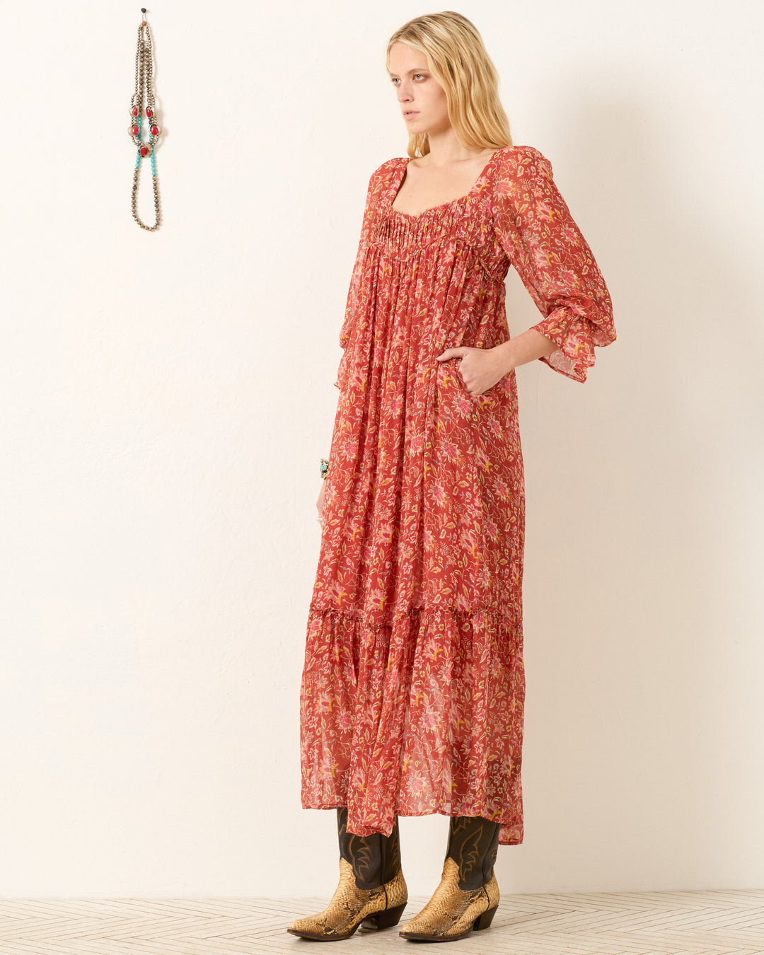 A woman in the Lilium Sunset Ember Dress, a red floral maxi with a flowy silhouette, pairs it with brown cowboy boots and stands sideways by a plain light wall, with necklaces displayed beside her.