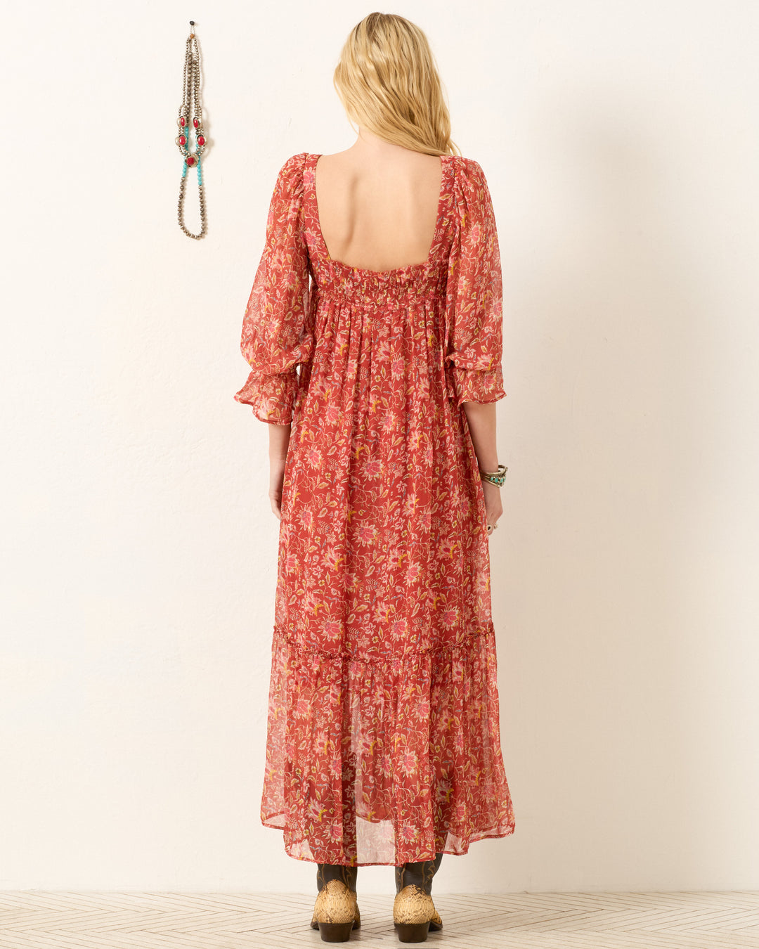 Woman with her back to the camera wears the Lilium Sunset Ember Dress, a red floral maxi with puffed sleeves and a flowy silhouette, paired with sandals. She stands before a light wall decorated with hanging beaded necklaces.