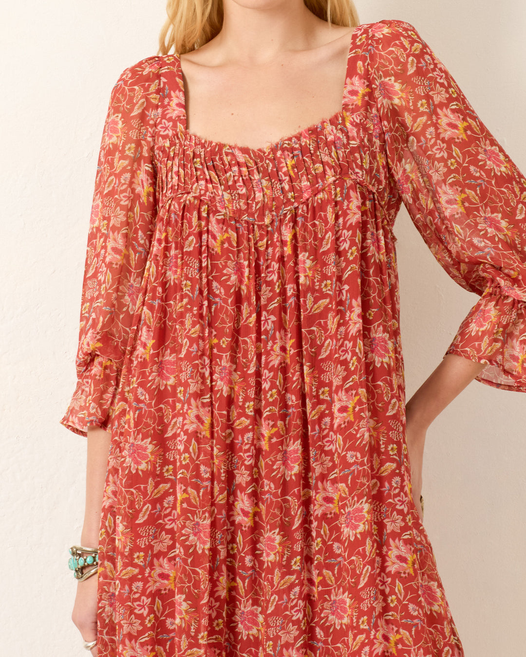 A woman wears the Lilium Sunset Ember Dress, a loose-fitting red floral maxi with long sleeves, a square neckline, gathered chest detailing, and a flowy silhouette.