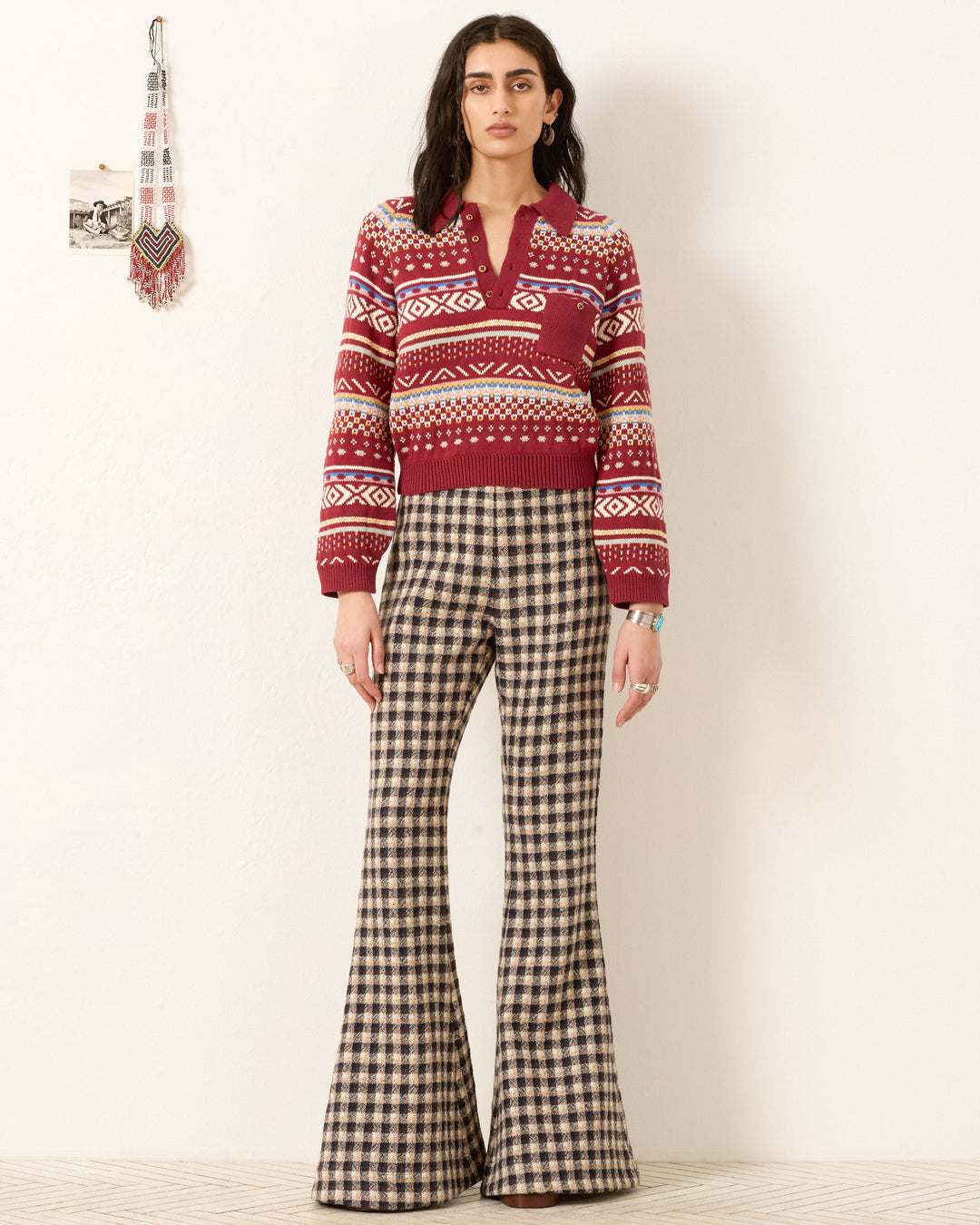 A woman stands against a plain wall wearing the vintage-inspired Liza Mesa Fair Isle Top and high-waisted, brown plaid flared pants.