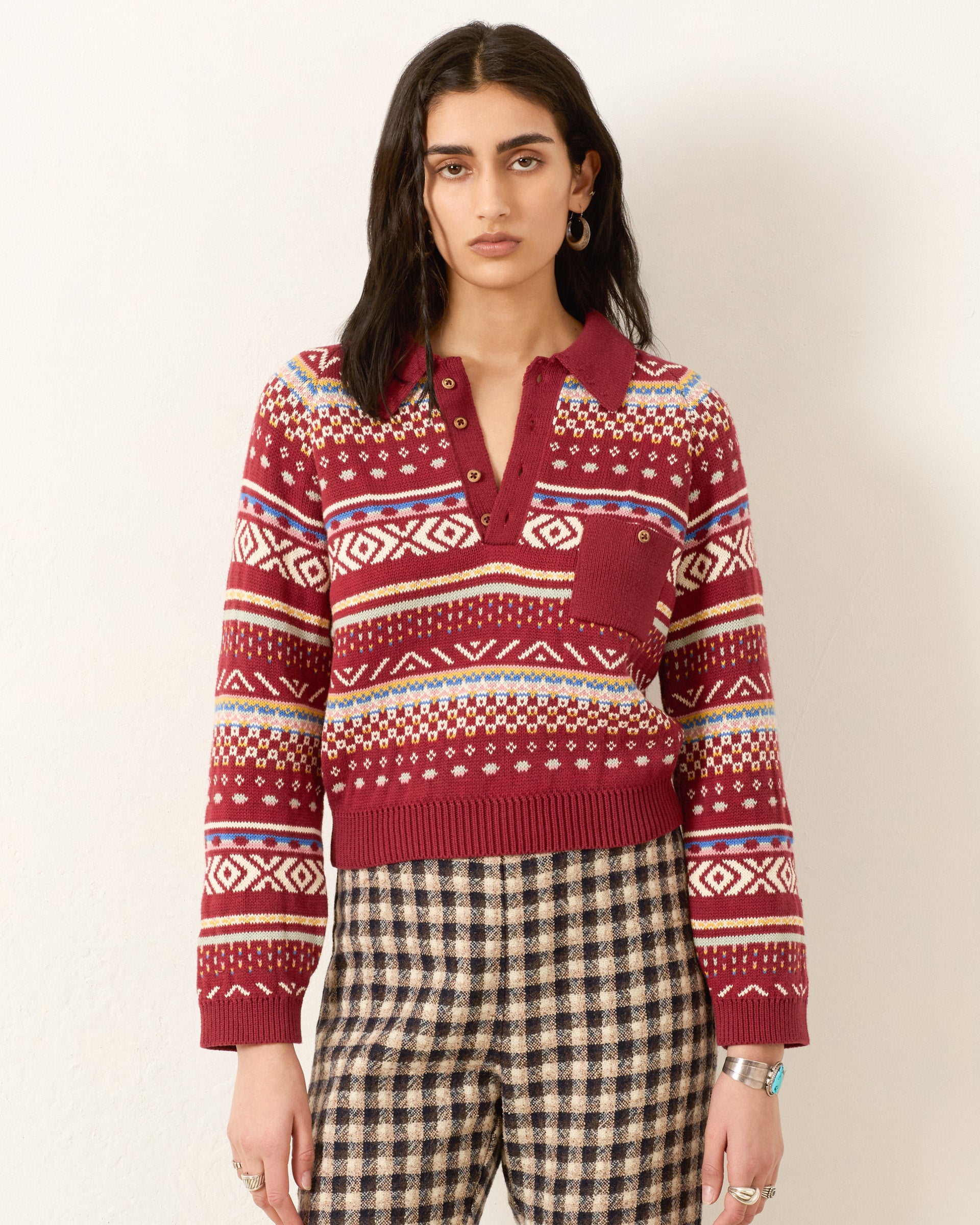 A person stands against a plain wall, wearing the Liza Mesa Fair Isle Top— a vintage-inspired red patterned cotton polo sweater with a partial button placket—paired with checkered pants.