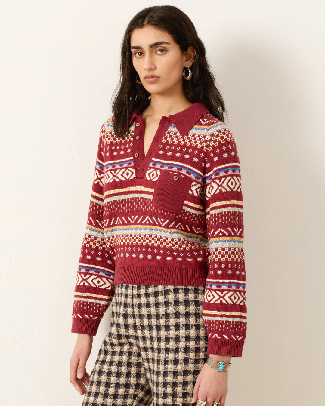 A woman stands against a plain wall wearing the Liza Mesa Fair Isle Top, a vintage-inspired sweater with a collar and button placket, paired with high-waisted plaid pants.
