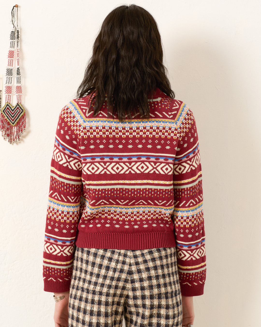 A person with wavy dark hair, seen from behind, wears the Liza Mesa Fair Isle Top—a vintage-inspired red patterned sweater—paired with plaid pants, standing before a white wall with a beaded wall hanging.