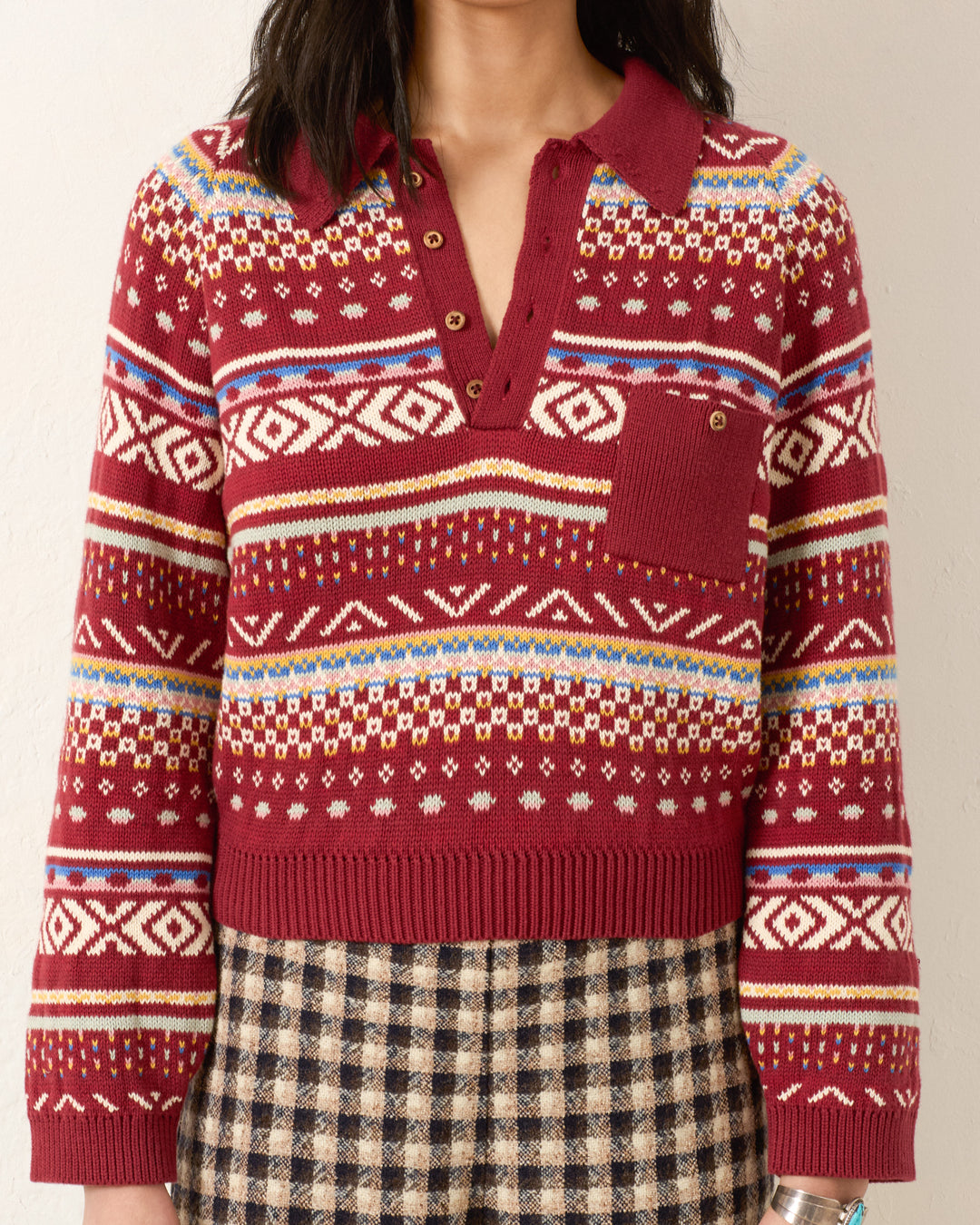 A person wearing the Liza Mesa Fair Isle Top, a vintage-inspired red patterned knit sweater with a collar and buttons, paired with a black and beige checkered skirt.