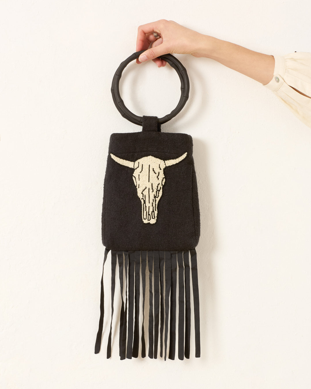 A hand holds the Lorien Cow Skull Beaded Bag, featuring a round handle, bold white cattle skull design, and long black fringe at the bottom.