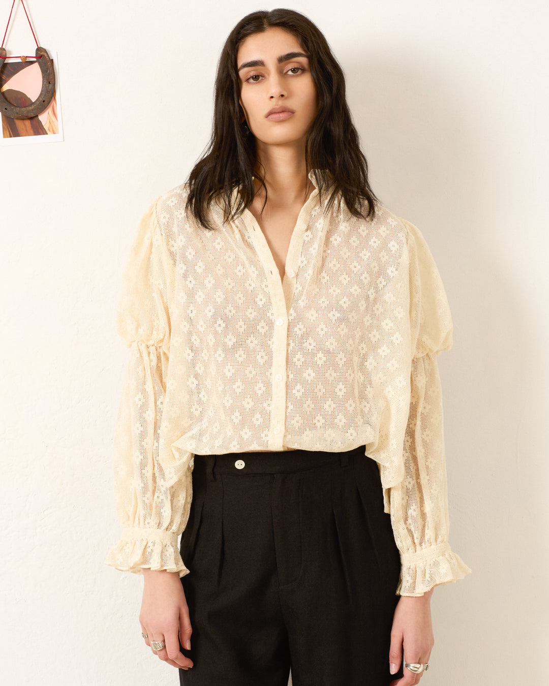 Person standing against a white wall, wearing the Mazie Antique Bone Lace Shirt with patterned ruffled cuffs in true ’70s dandy style, paired with black high-waisted trousers.