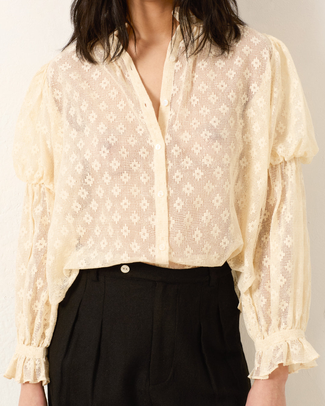 Wearing the Mazie Antique Bone Lace Shirt, a sheer cream cotton top with long sleeves, paired with black high-waisted pants for a ’70s dandy-inspired look.
