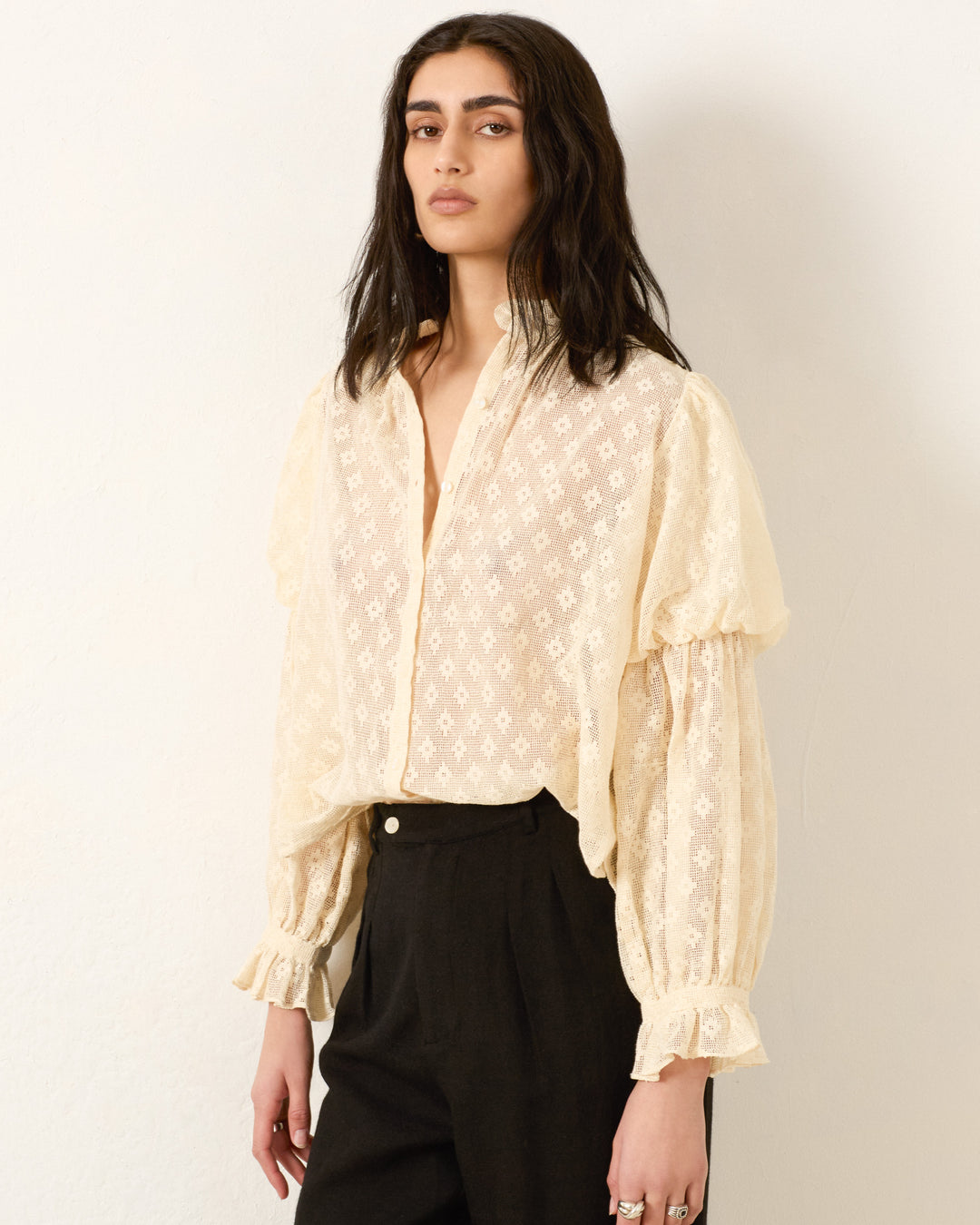 A person wears the Mazie Antique Bone Lace Shirt, featuring cream-colored lace and puffed sleeves in a '70s dandy style, paired with black high-waisted pants against a plain light background.