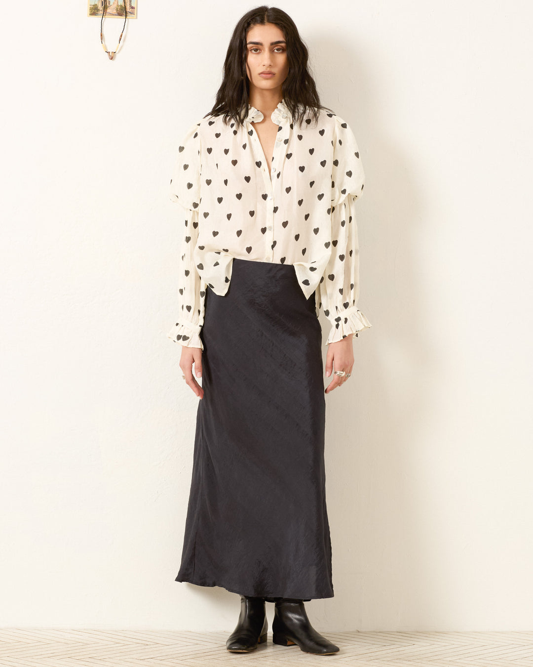 A person stands against a plain wall, wearing the Mazie Onyx Heart Silk Shirt with black heart prints, paired with a long black skirt and black ankle boots.