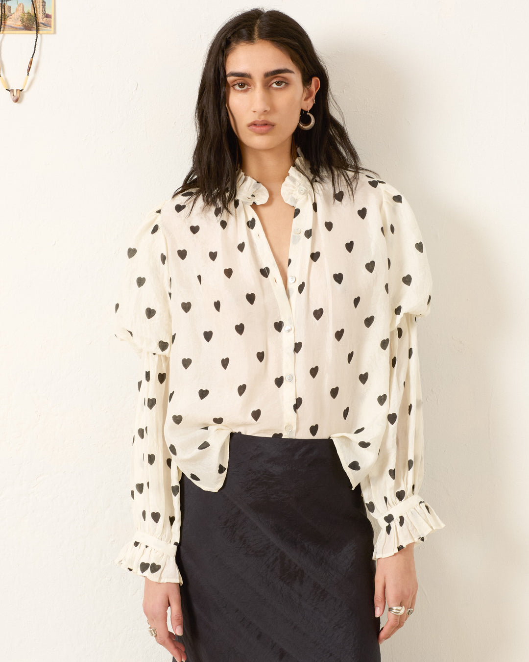 A person stands against a light wall, wearing the Mazie Onyx Heart Silk Shirt—a cream hand-block printed blouse with black heart pattern and ruffled 70s-style cuffs—paired with a black skirt.