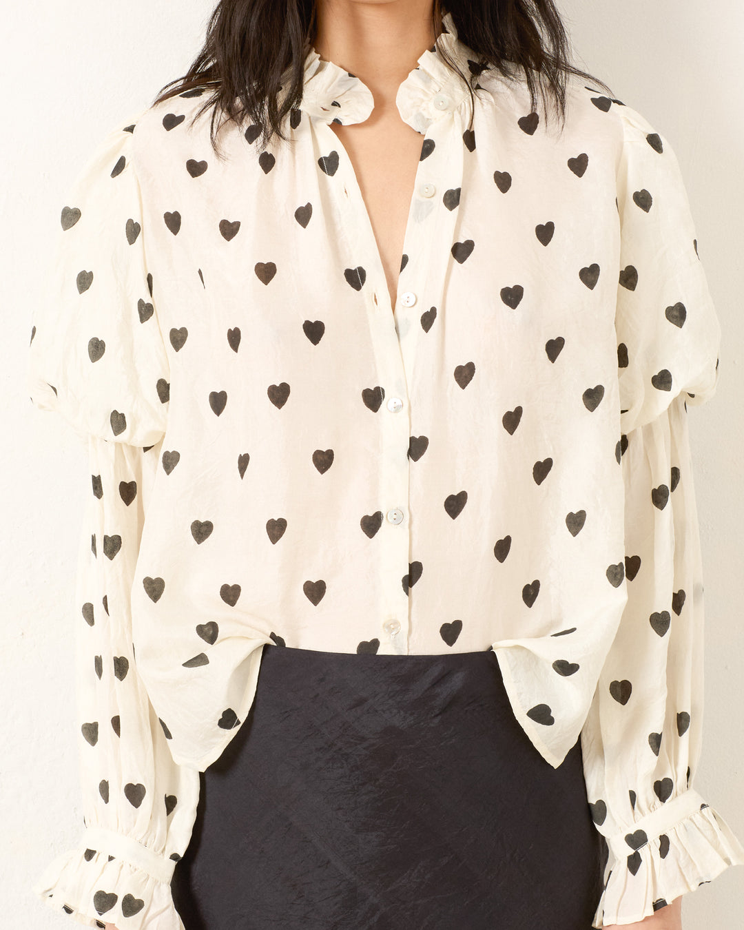 Wearing the Mazie Onyx Heart Silk Shirt—a white hand-block printed blouse with black heart prints and puffed sleeves—paired with a black skirt for a playful ’70s dandy style.