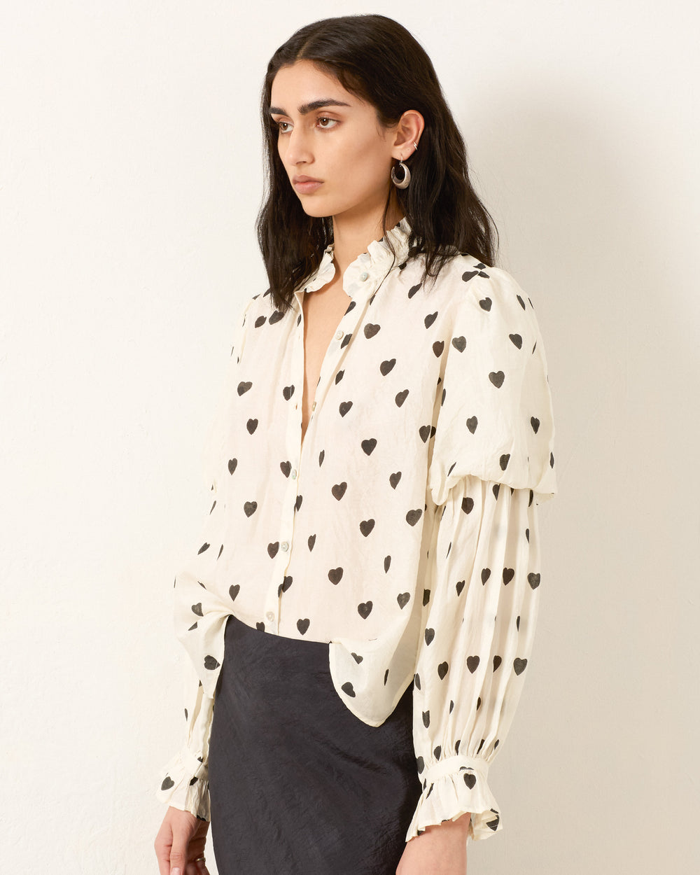 A woman stands against a plain wall wearing the Mazie Onyx Heart Silk Shirt with black heart patterns and a black skirt, evoking subtle 70s dandy style.