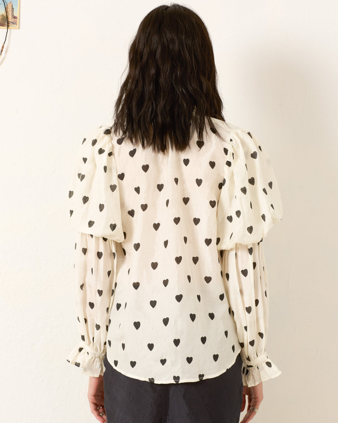 A person with wavy dark hair is seen from behind wearing the Mazie Onyx Heart Silk Shirt, a 70s-style white blouse with black heart patterns and puffed sleeves, set against a plain light wall.