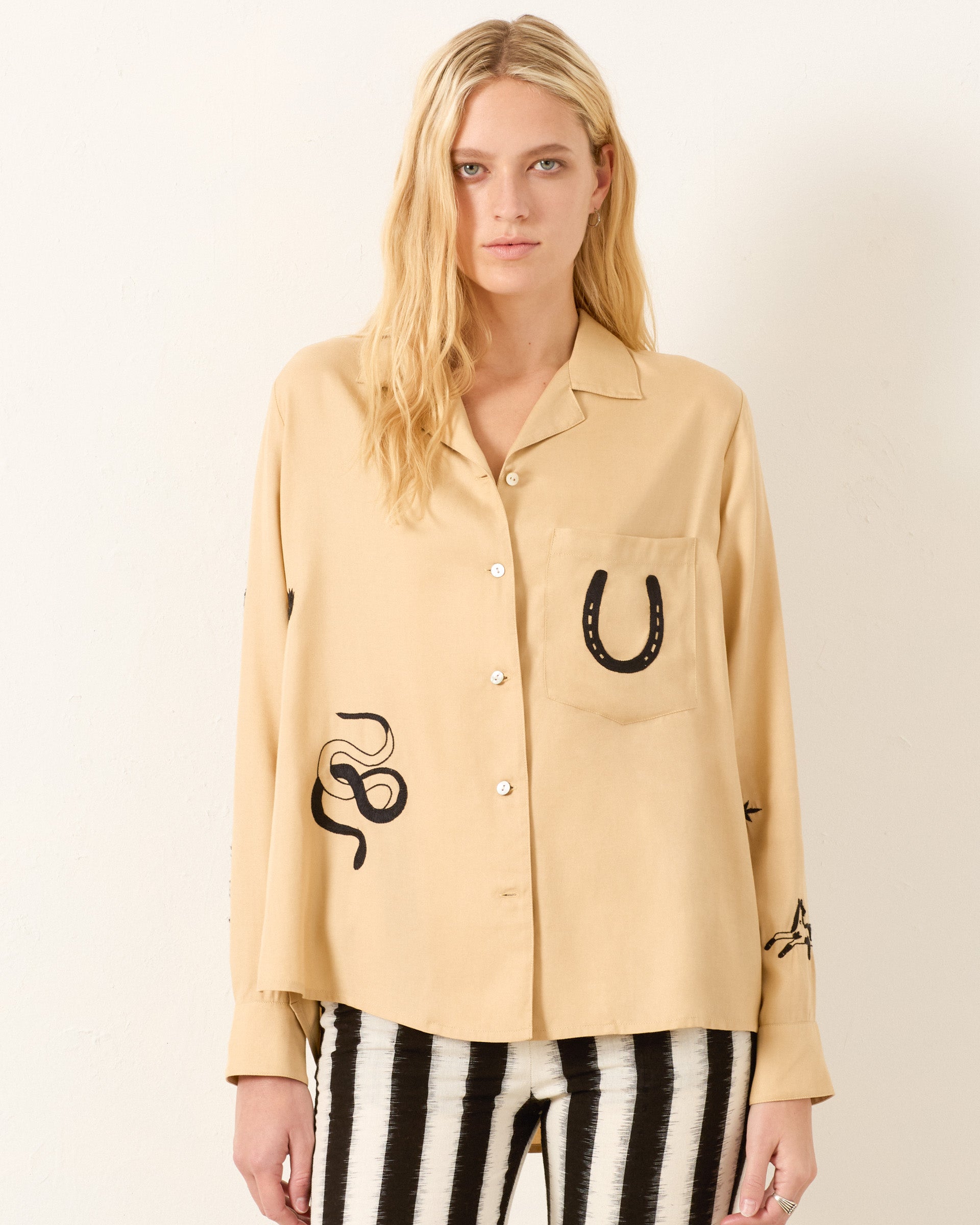 A woman wears the Patti Desert Tattoo Shirt, a beige hand-embroidered Western-inspired top with black designs like a horseshoe and snake, paired with black and white striped pants, standing against a plain light background.