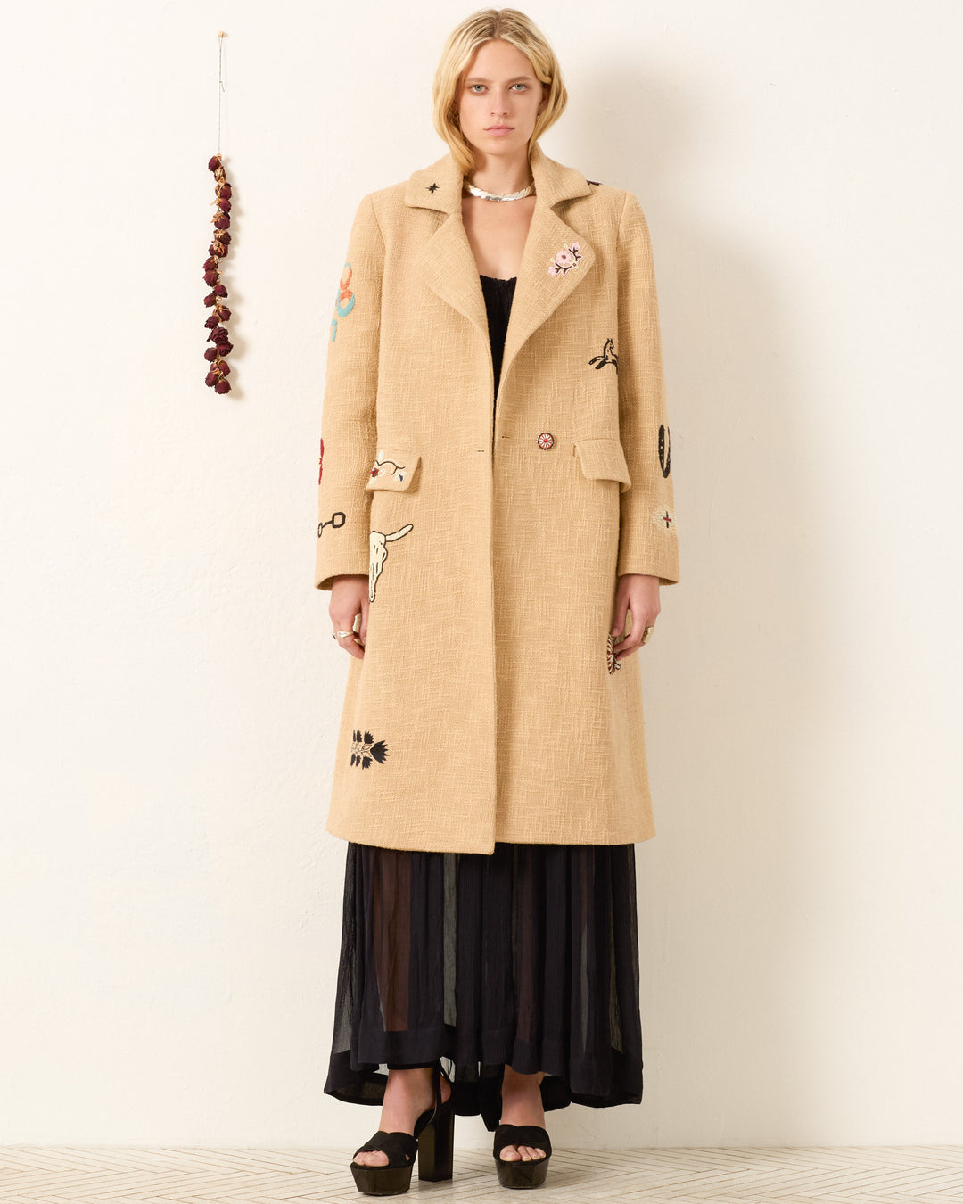 A woman faces forward wearing the Petunia Desert Tattoo Coat, a beige Khadi cotton piece with beaded embroidery, over a long black dress and platform heels. Behind her, a string of dried items hangs on the wall.