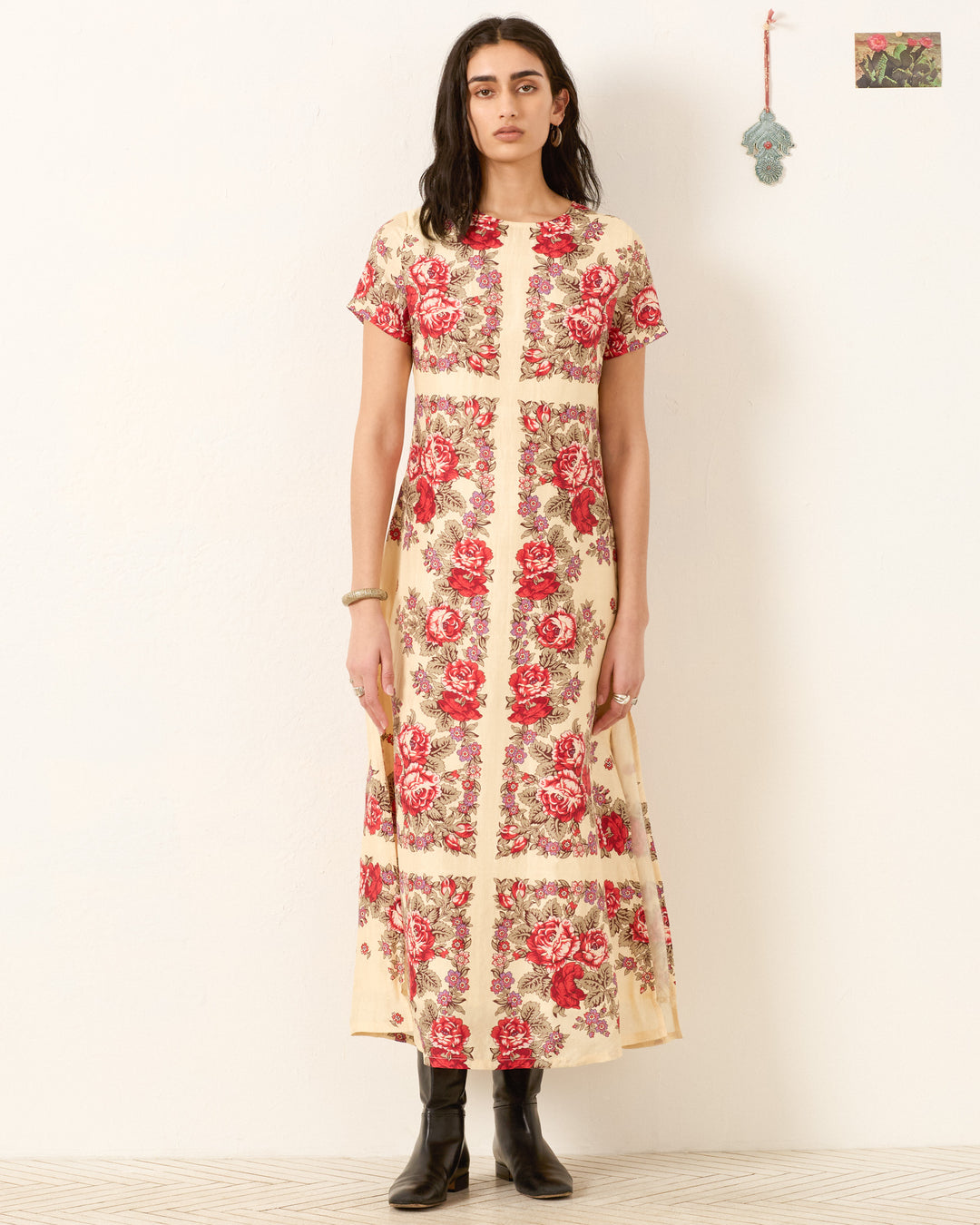 A woman stands against a white wall wearing the Preeti Rosebush Silk Dress, a cream-colored short-sleeve A-line maxi with red and pink flowers, paired with black boots and minimal jewelry.