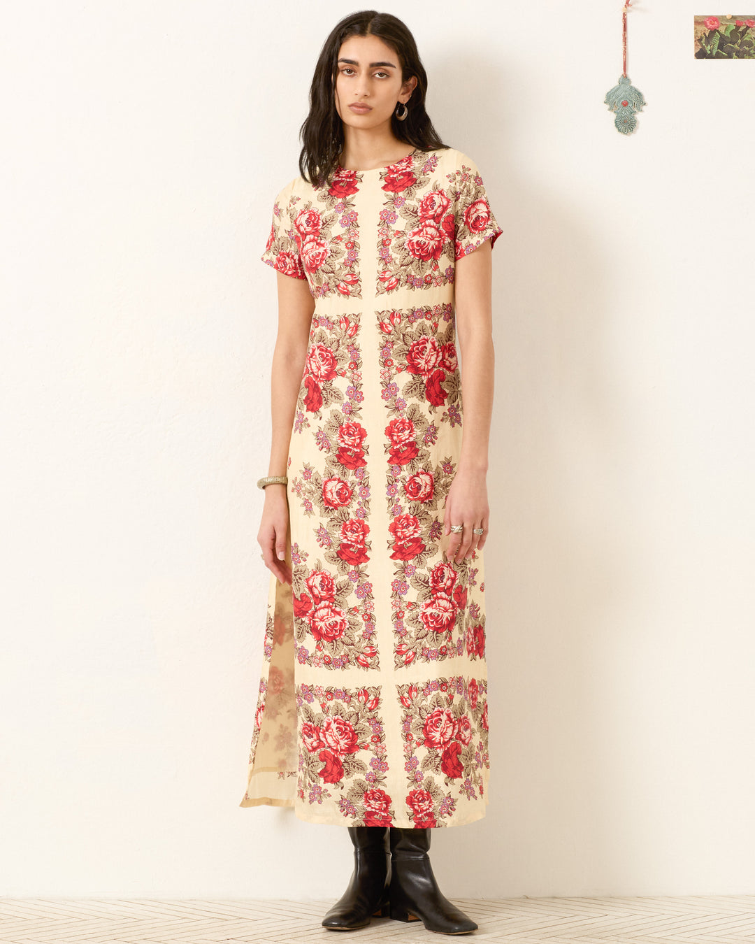 A person stands against a white wall wearing the Preeti Rosebush Silk Dress, a cream-colored A-line maxi with red floral patterns, paired with black boots and minimal jewelry.