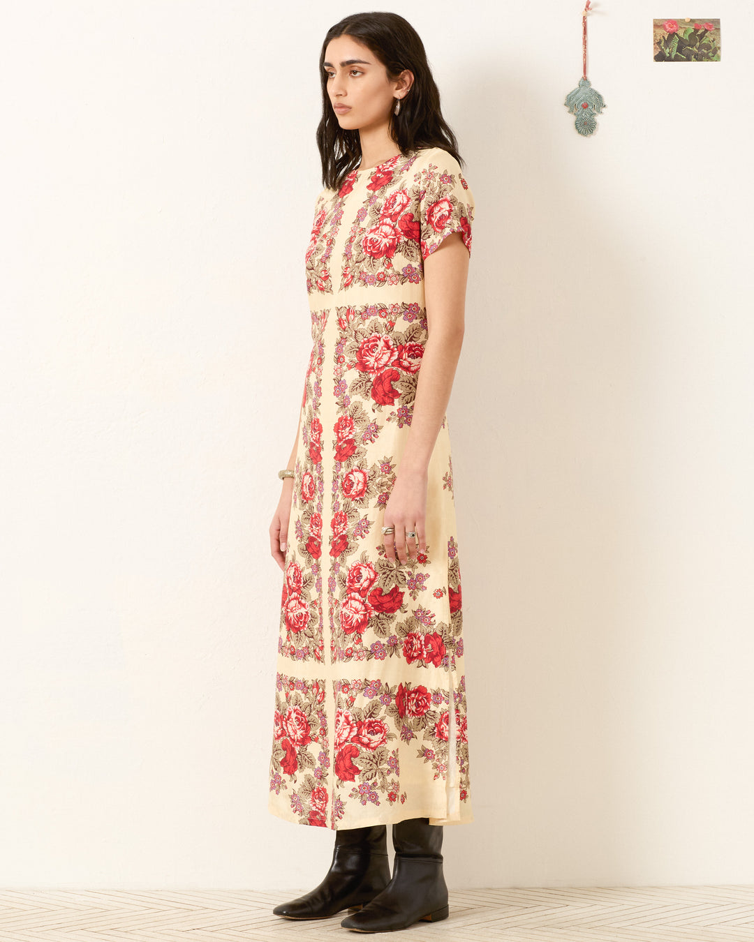 A person stands sideways in the Preeti Rosebush Silk Dress, a cream hand-loomed silk piece with a red floral pattern, paired with black boots against a plain light background.