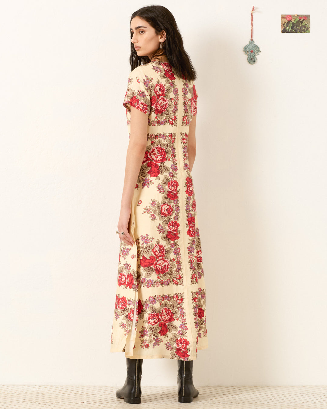 Woman stands with her back to the camera in the Preeti Rosebush Silk Dress, an A-line maxi with red floral prints, paired with black boots against a plain backdrop featuring a small wall decoration.