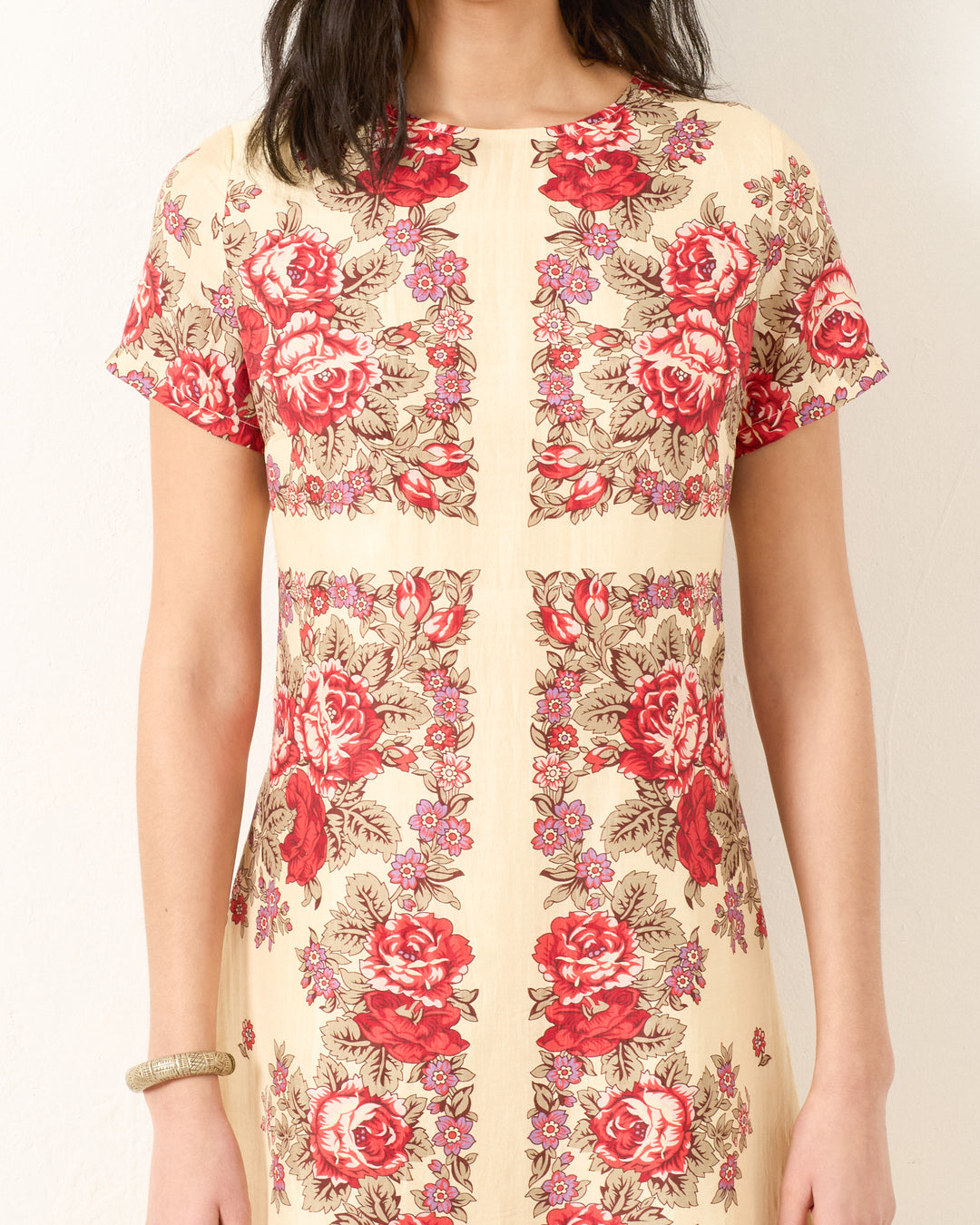 Someone wearing the Preeti Rosebush Silk Dress, featuring short sleeves and a cream base with a red rose motif, standing against a plain light-colored wall.