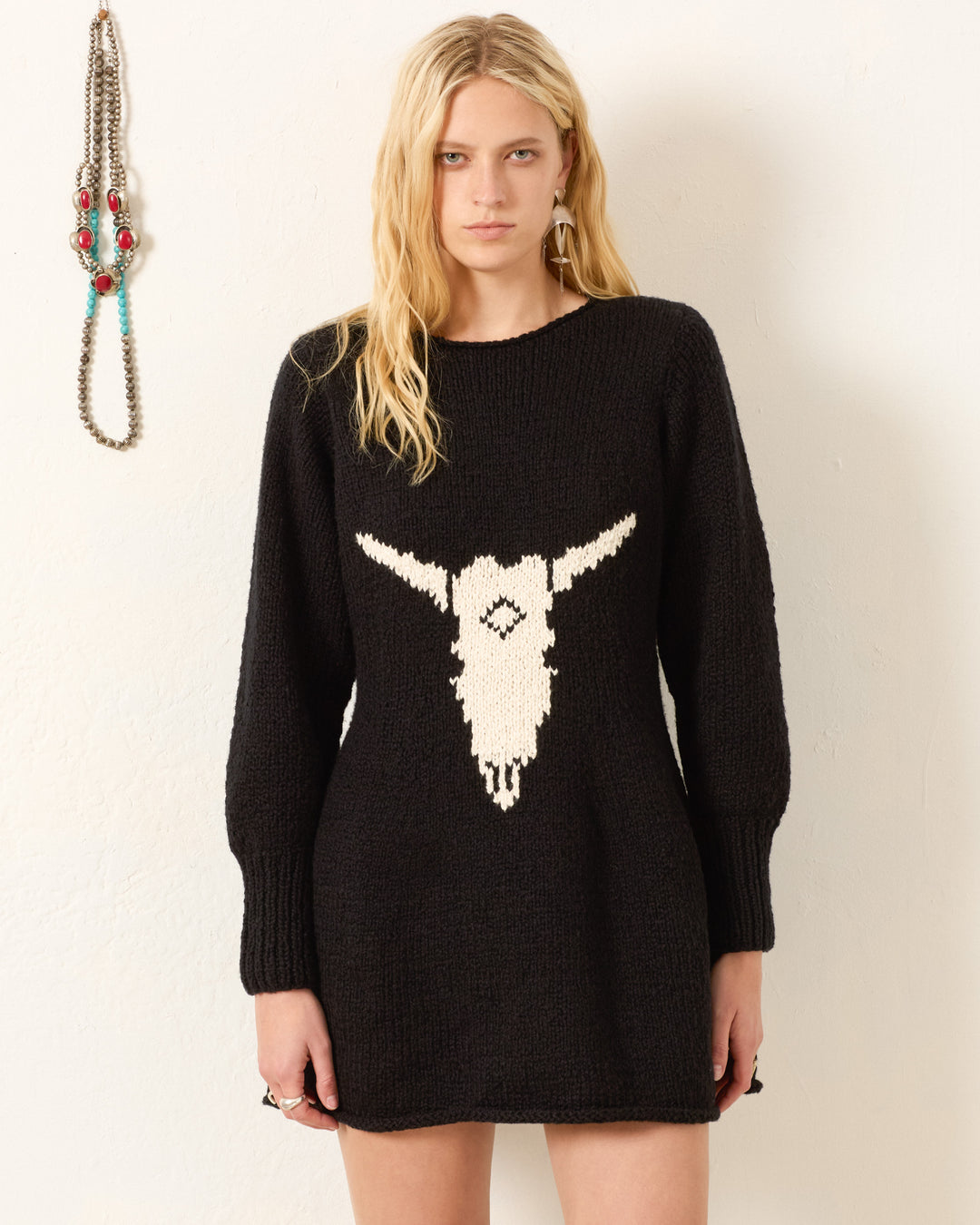 A woman stands against a white wall wearing the Renny Cow Skull Dress, a hand-knitted black sweater dress featuring a white animal skull design; beaded jewelry is displayed on the wall beside her.
