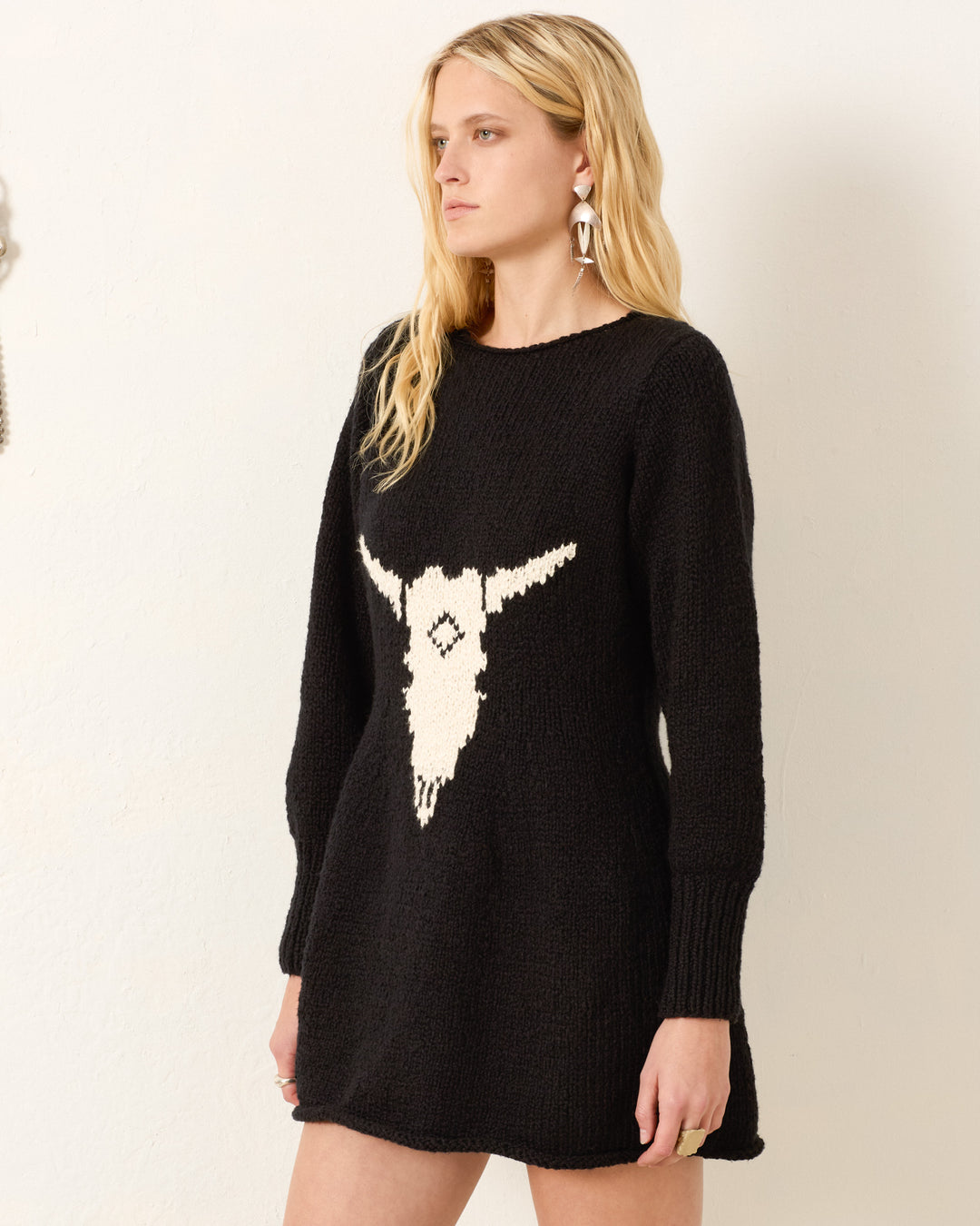 A woman with long blonde hair models the Renny Cow Skull Dress, a black sweater dress with a bold white skull graphic on the front, standing before a plain light background.
