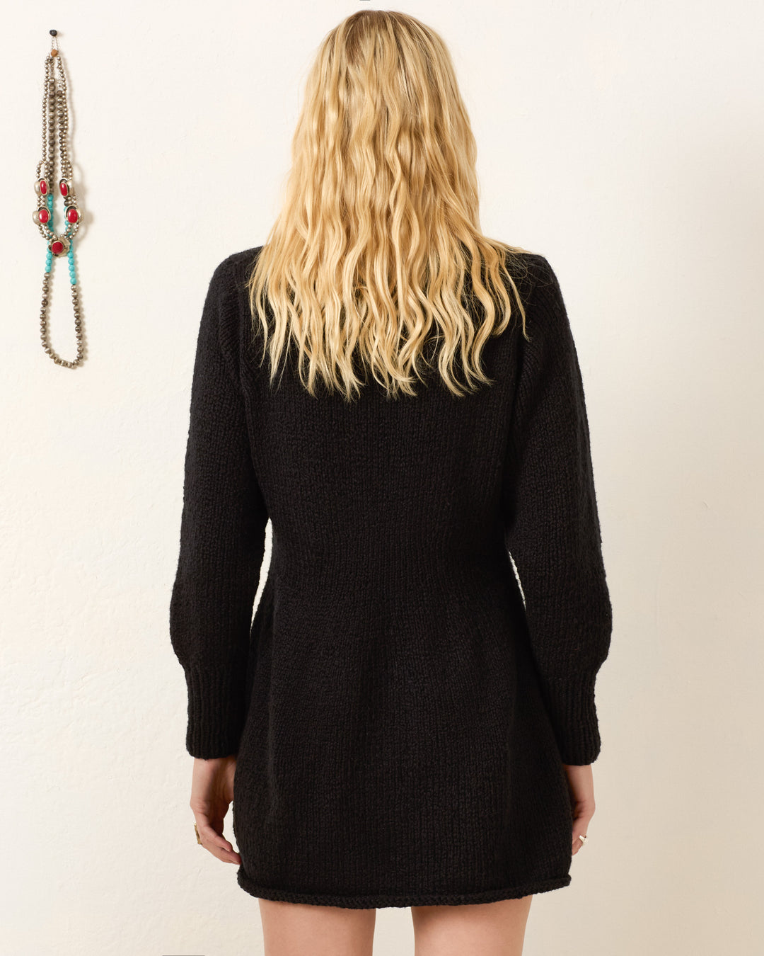 A person with long, wavy blonde hair is photographed from behind wearing the Renny Cow Skull Dress, a black, long-sleeve sweater dress. A beaded necklace hangs on the wall to the left.